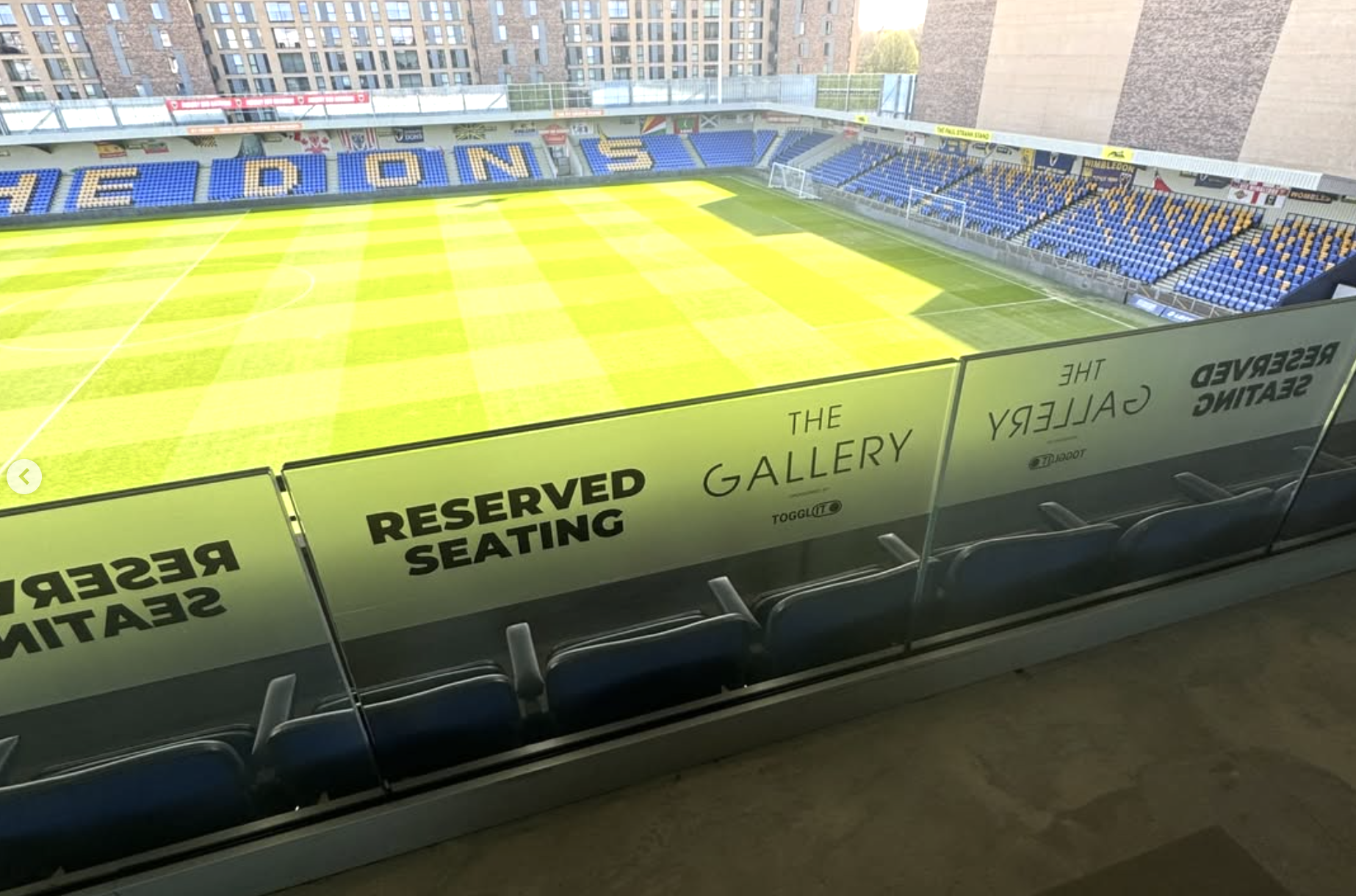 Empty afc wimbledon football ground  seats with reserved seating signs, overlooking a green field with goalposts, surrounded by high-rise buildings in the background.