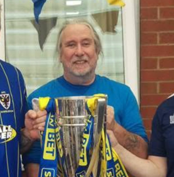 A man with gray hair and a beard, director of WISH John Lynch, holding a silver trophy with yellow and blue ribbons, smiling in front of a brick wall and a glass window.