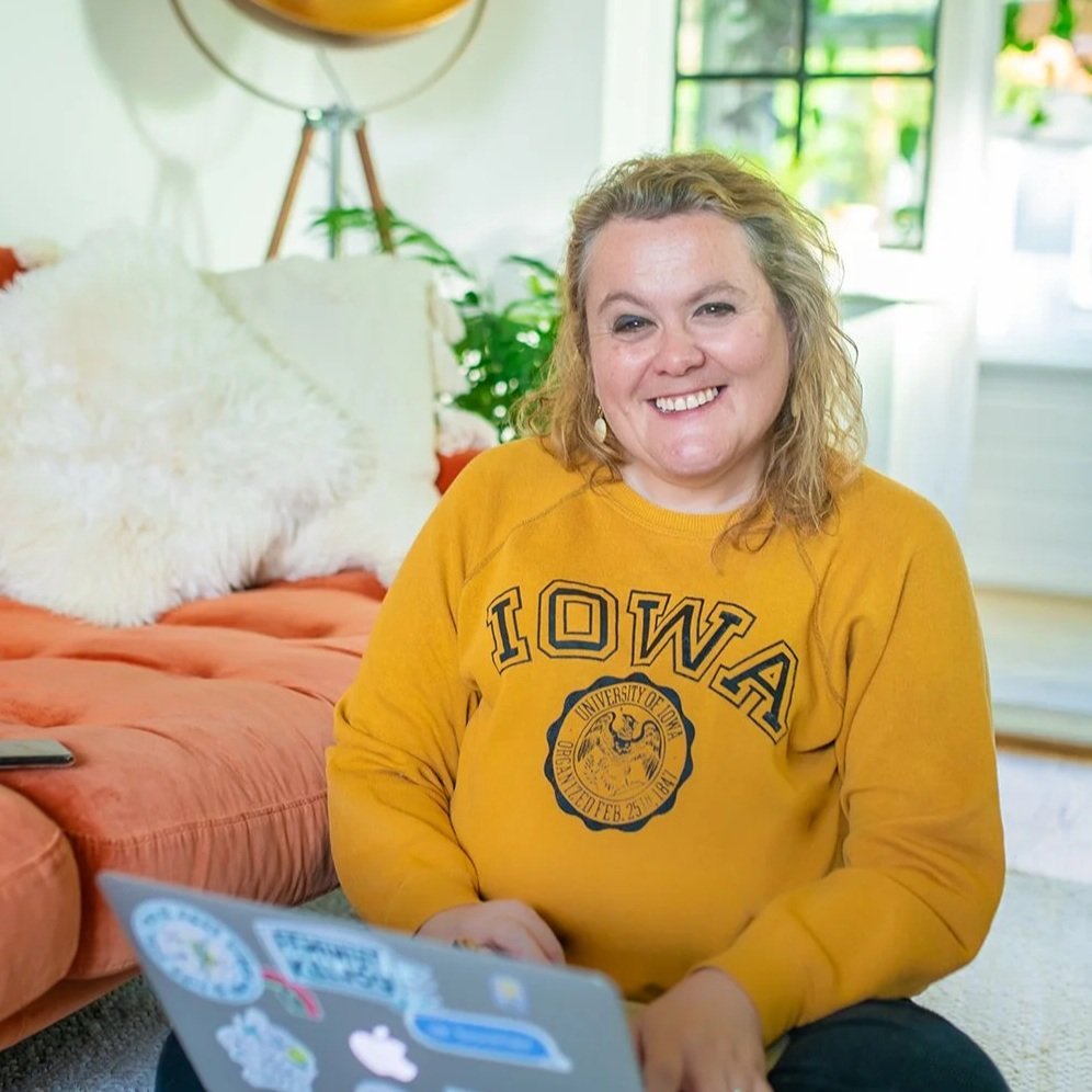 Jude Turner Creative, a Woman smiling while sitting indoors with laptop, wearing a yellow Iowa sweatshirt, in a cozy room with a sofa, cushions, and plants.