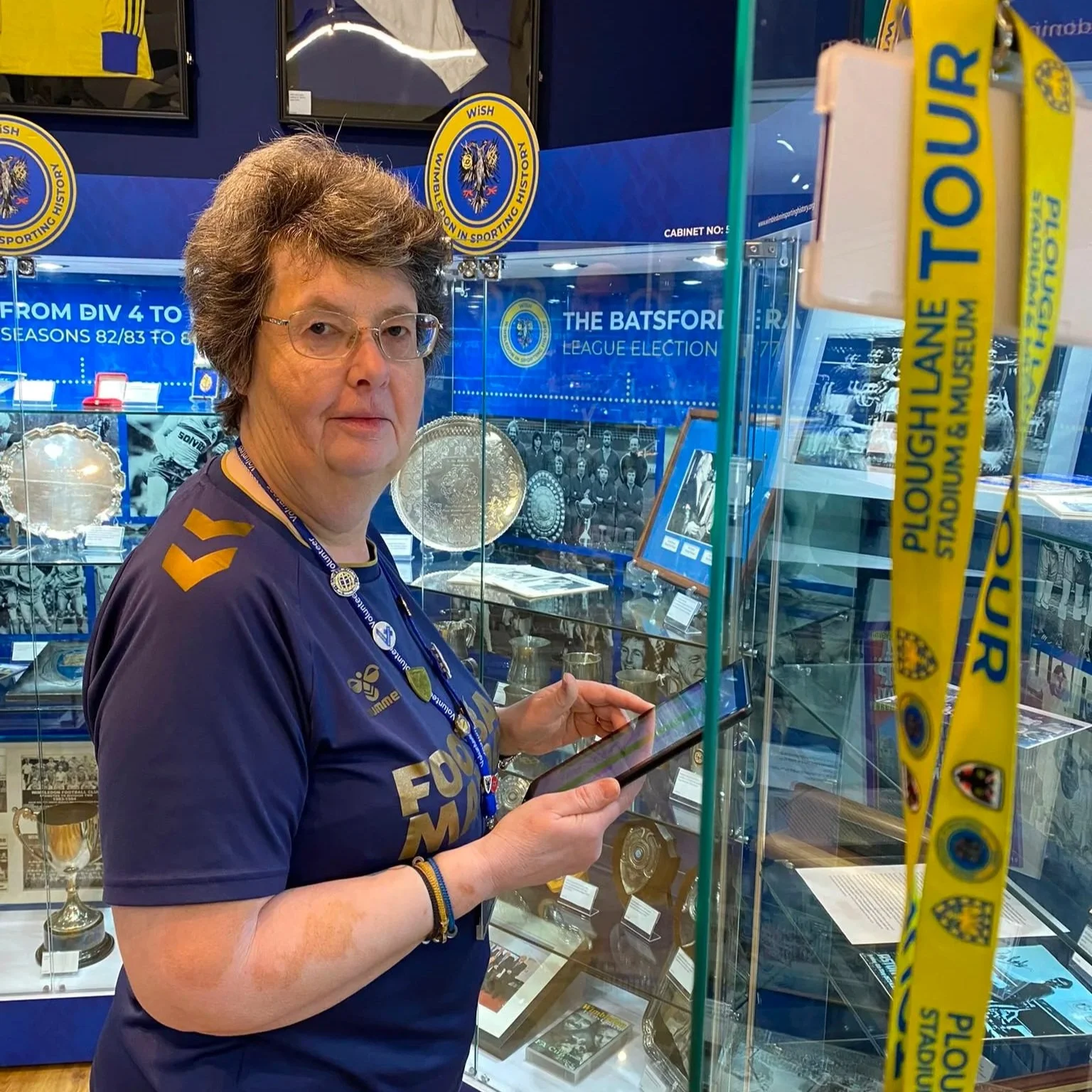 A woman with glasses wearing a blue sports jersey and a lanyard, standing in front of a display case filled with sports memorabilia, in a museum dedicated to the history of the national football league.