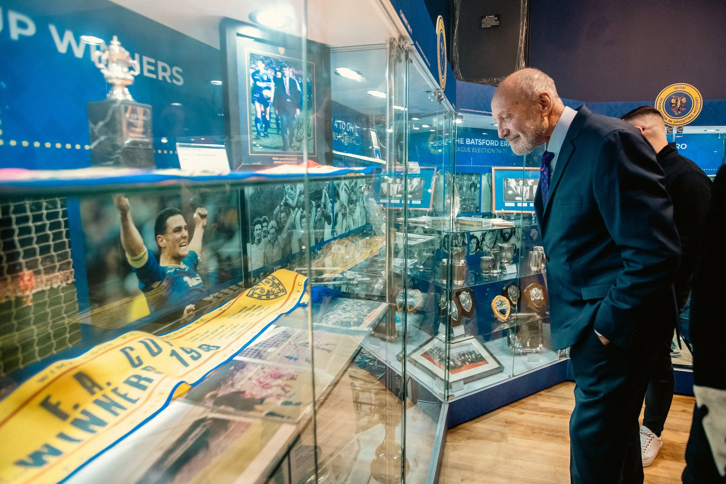 A display case with sports memorabilia, including a photograph of a football player celebrating and various items related to AFC Wimbledon Football Club, with a man in a suit, Dickie Guy,  looking at it.