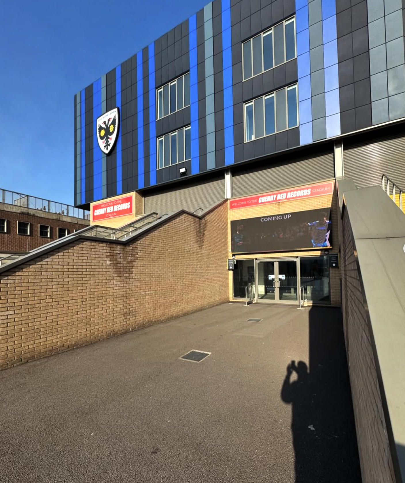 Exterior view of  afc wimbledon ground  with a sign welcoming to Cherry Red Records Stadium, and the crest of a football club on the building wall, under a clear blue sky.