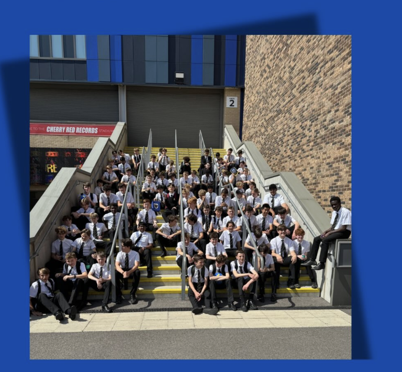 Group of young students in uniform sitting and standing on outdoor staircases on a stadium tour. They are wearing school uniform. The background features a brick wall, stairs, and a storefront with a sign "Cherry Red Records." 