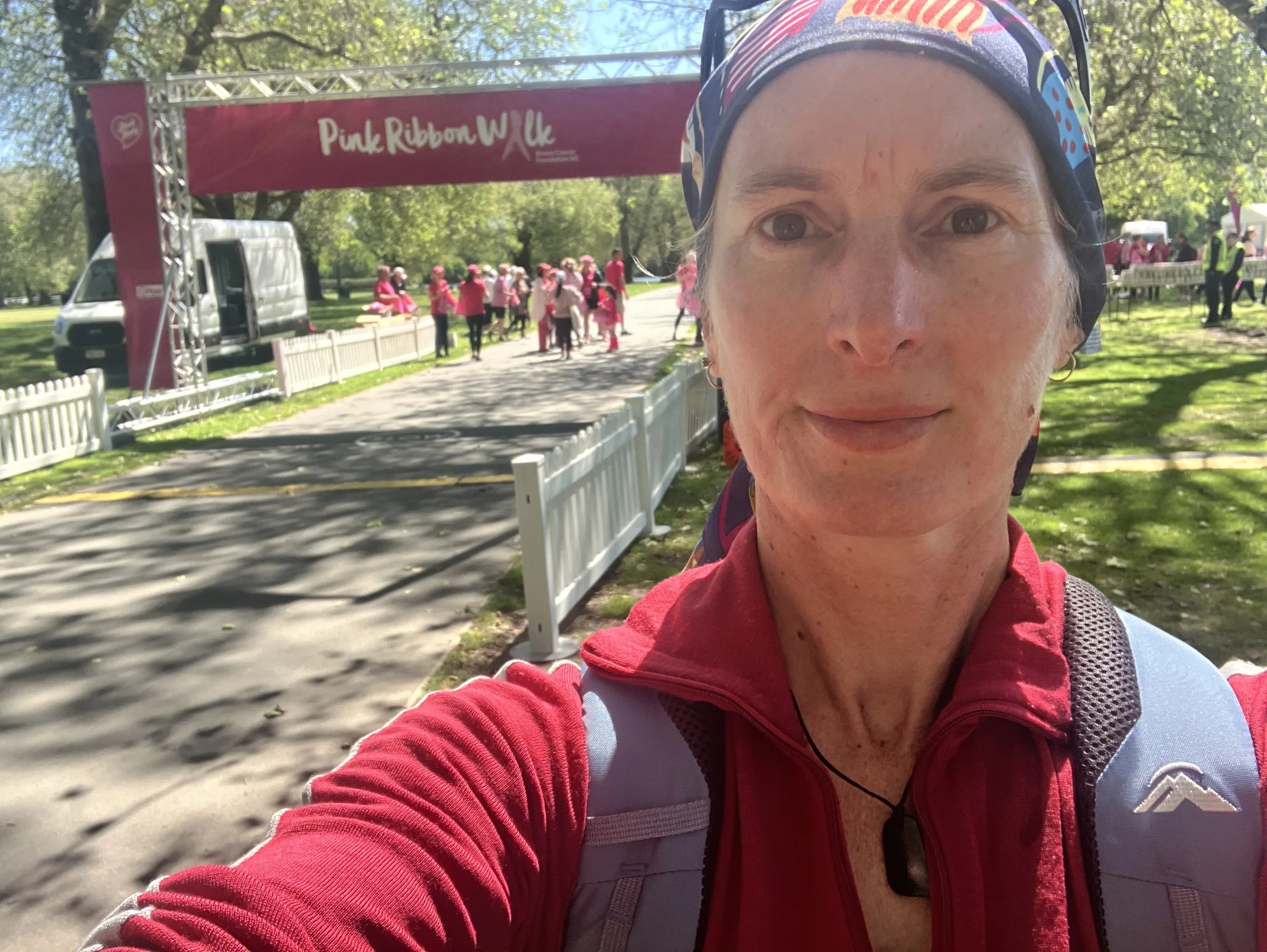 Close-up of a woman wearing a colorful headband and a red jacket, taking a selfie at the Pink Ribbon Walk event. In the background, people dressed in pink are gathering near a pink banner that reads 'Pink Ribbon Walk' in a park with green trees.