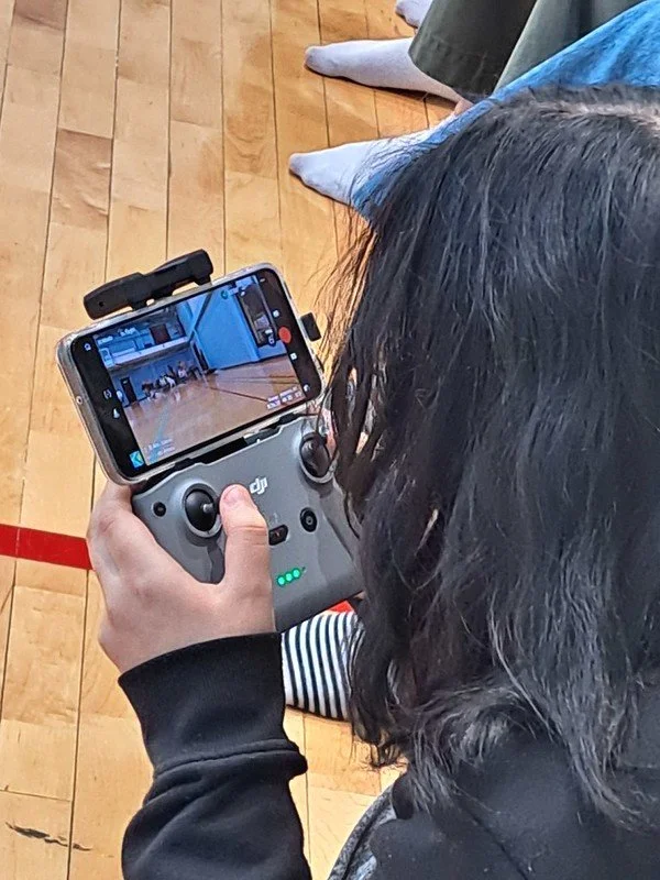 Person with dark hair operating a drone remote control with a smartphone mounted on top, capturing images or video of an indoor gymnasium with wooden floors and people in the background.