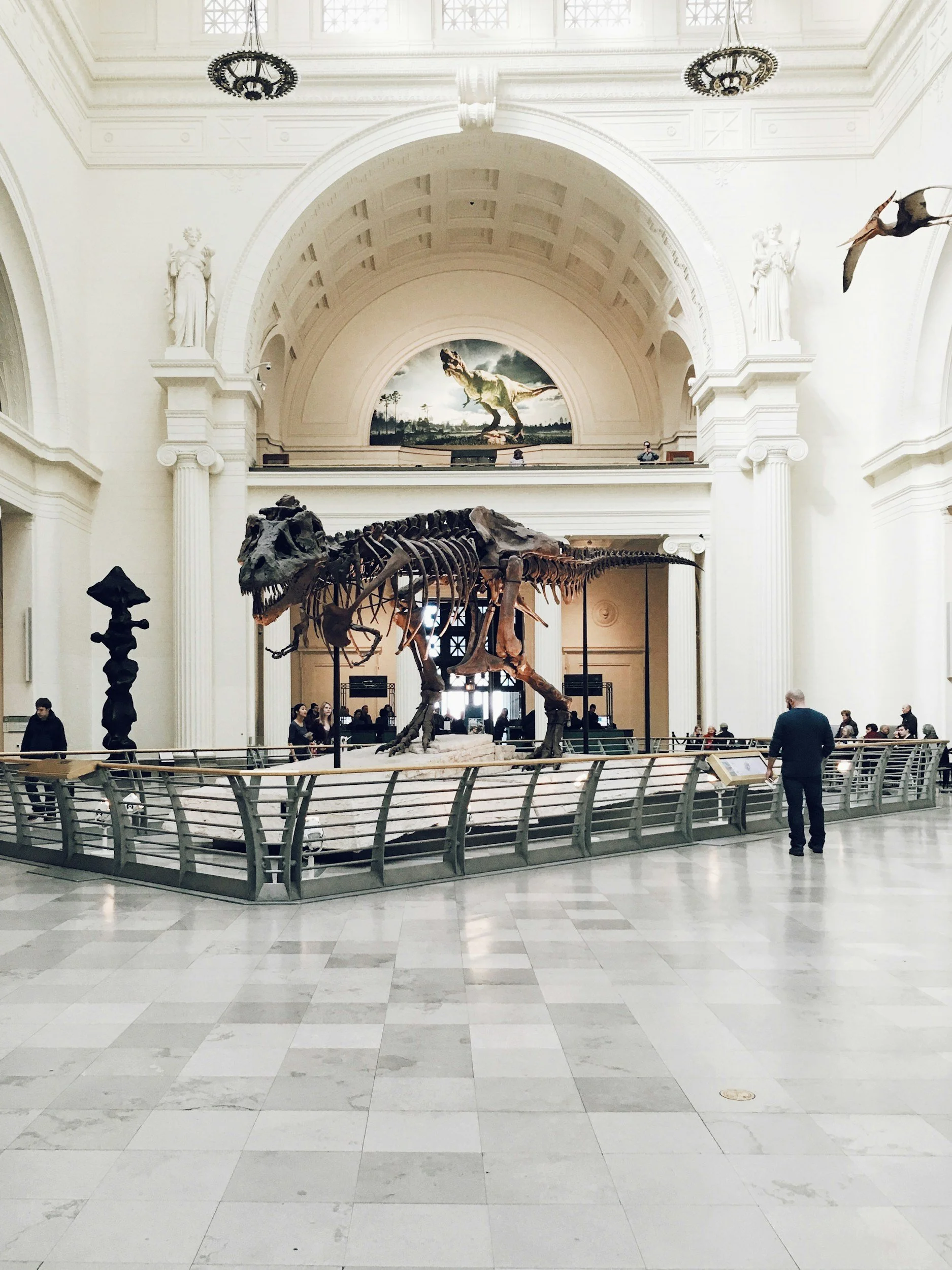 Hall with dinosaur skeleton exhibit, white walls, ornate ceiling, statue sculptures, and visitors observing the display.
