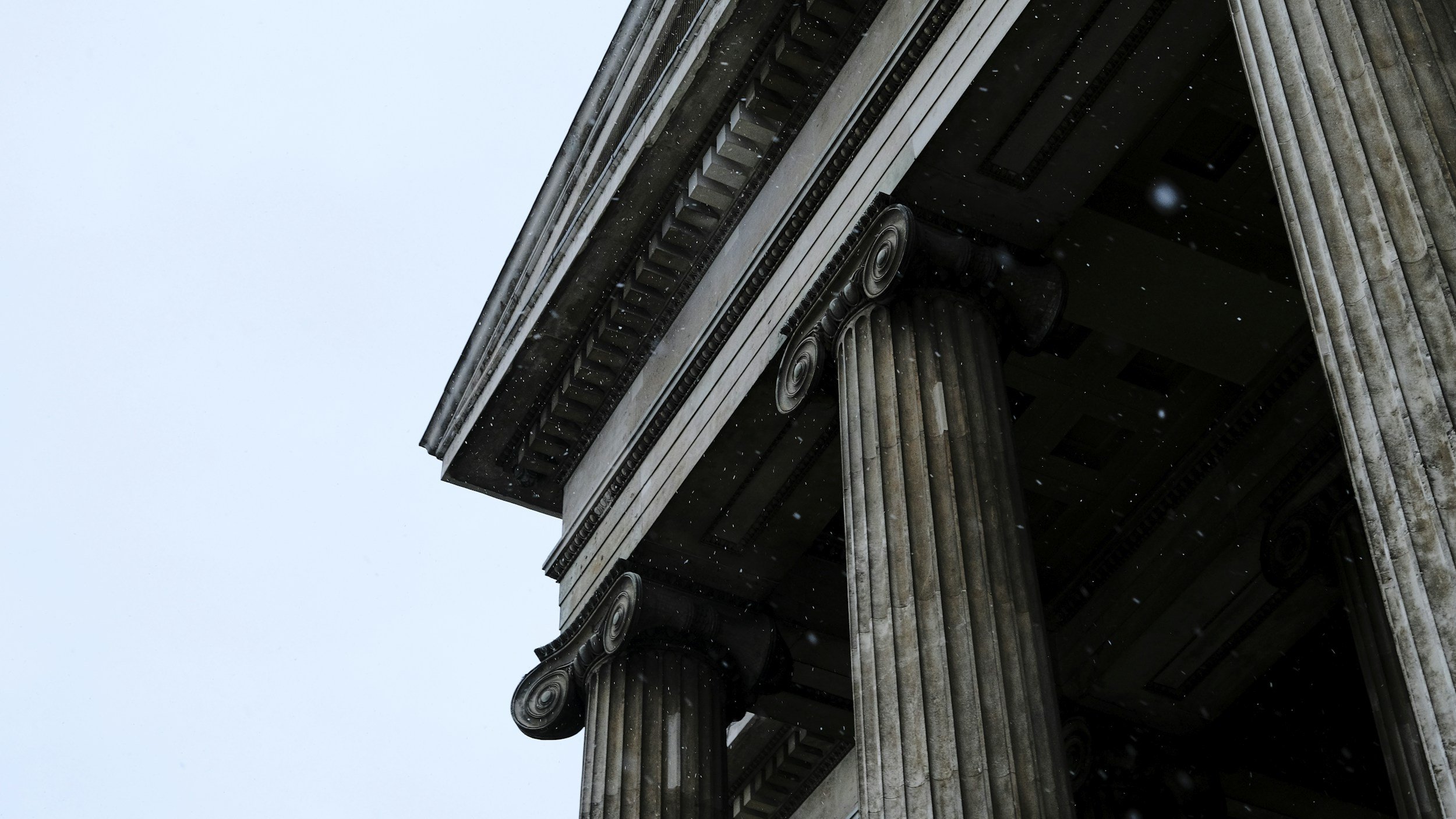 Close-up view of a building with classical architectural details, including Corinthian columns and decorative friezes, taken from below against a cloudy sky.