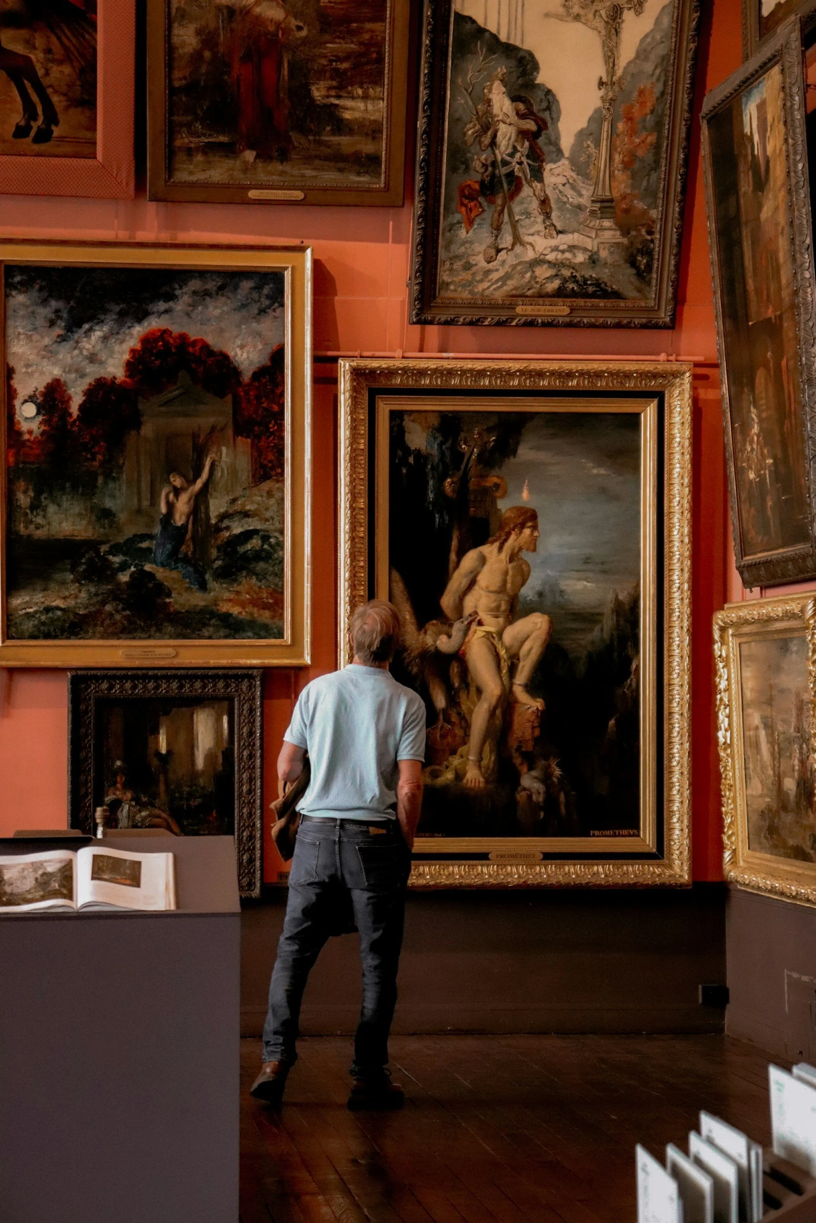 A man observing framed paintings in an art gallery or museum with a dark wooden floor and pinkish wall.