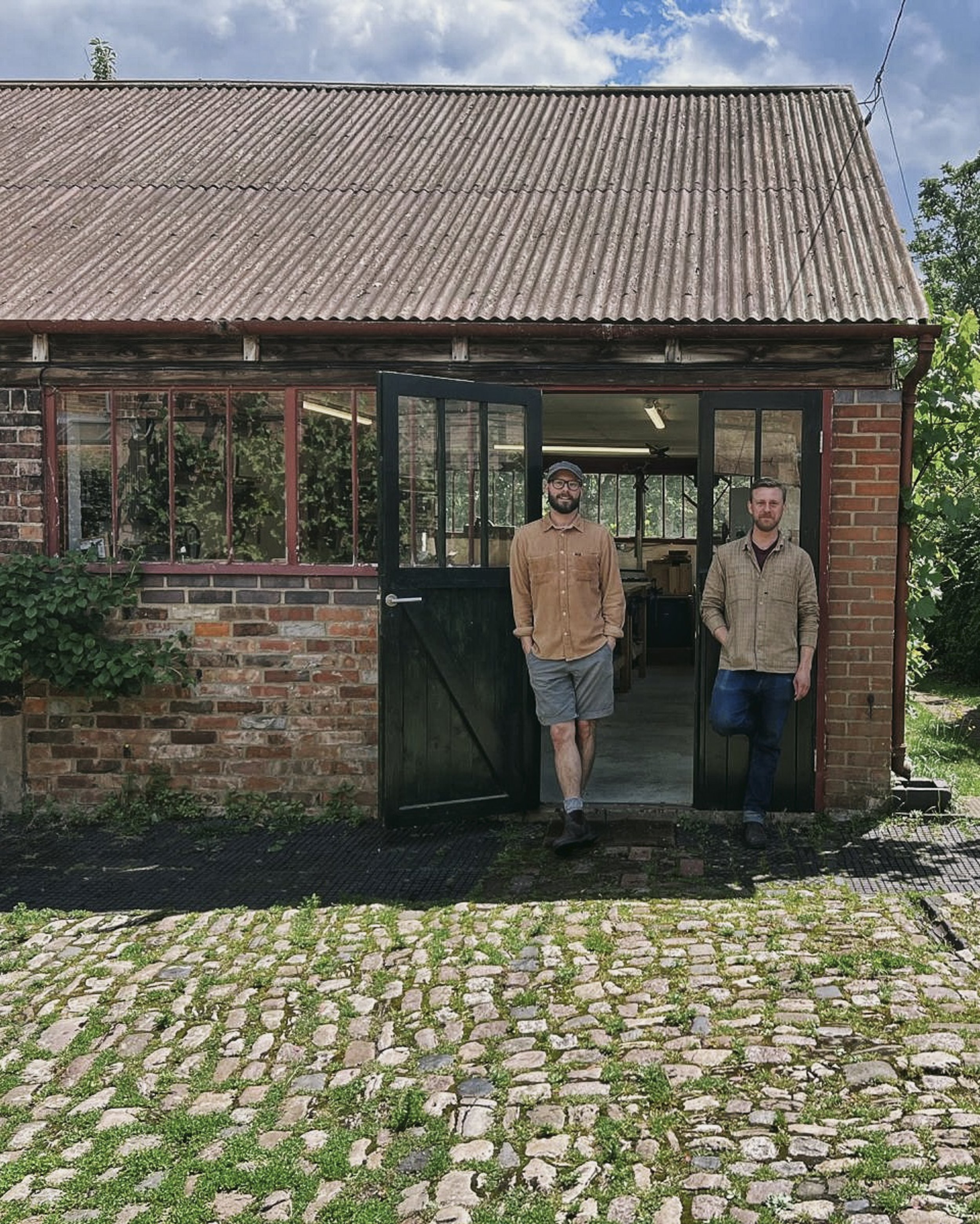 Two men standing at the open entrance of a brick building with large windows, under a sloped corrugated metal roof, with a cobblestone pathway and green grass outside.