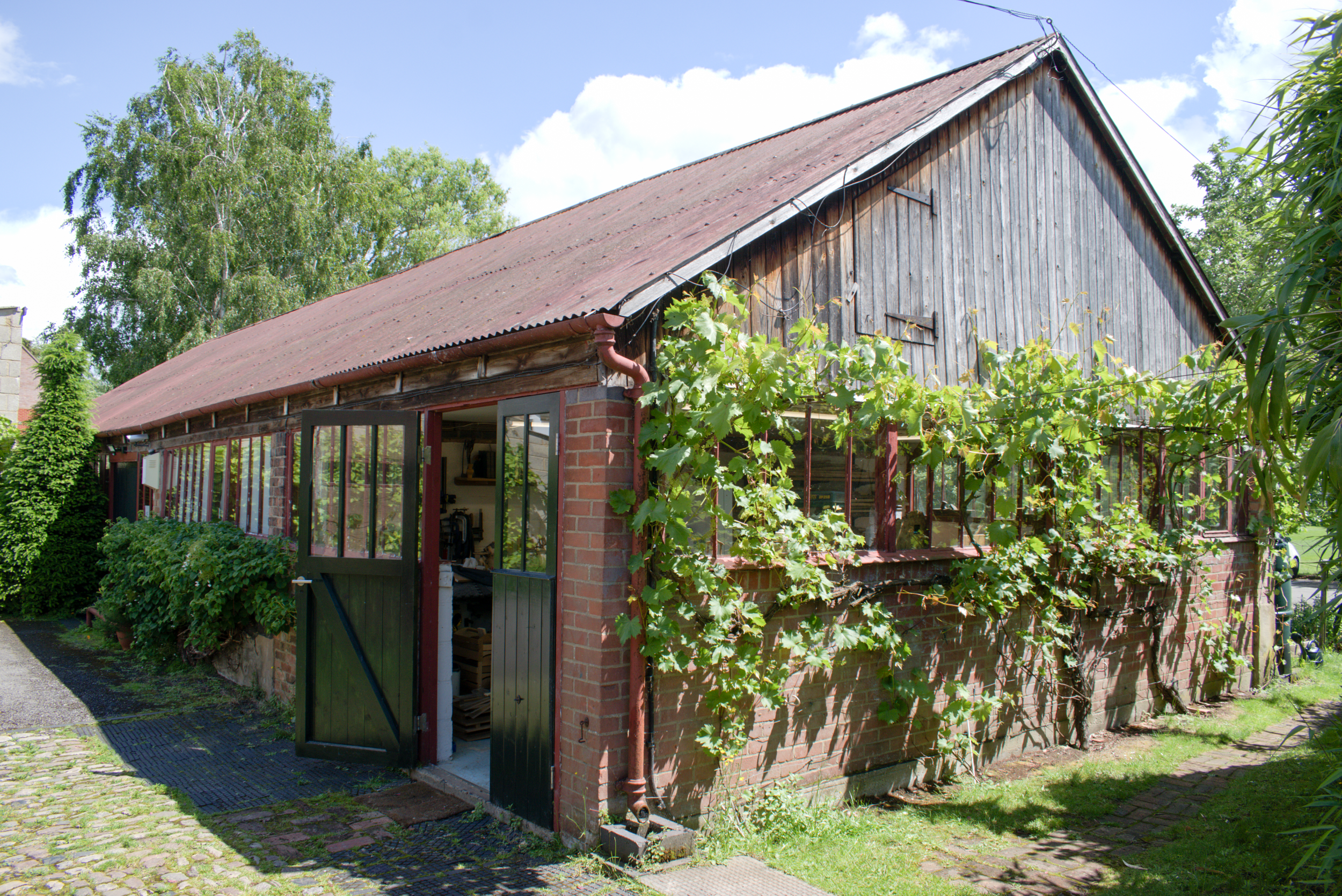 A rustic barn with wooden siding and a rusty metal roof, surrounded by green trees and plants, with a cobblestone pathway in front.