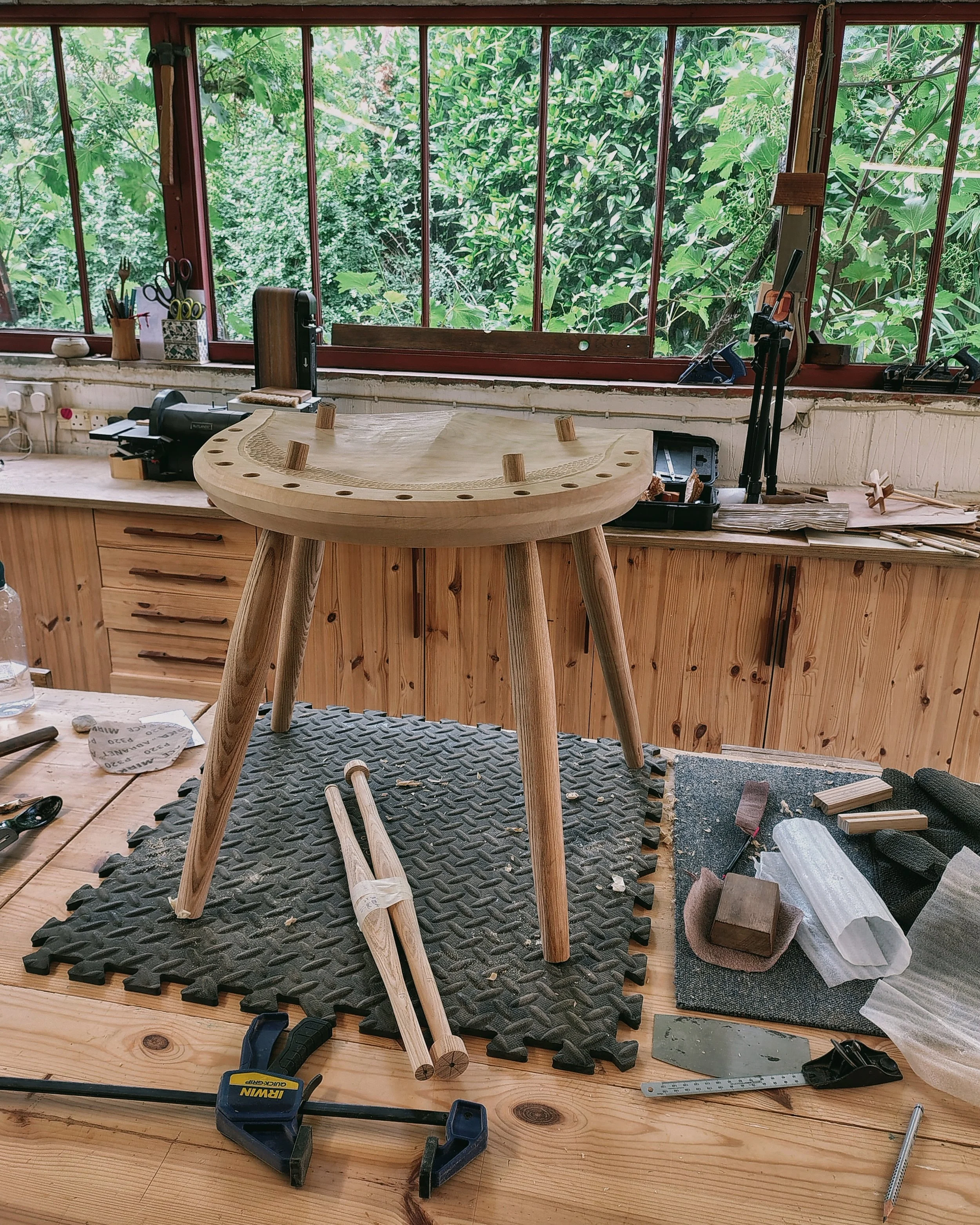 A woodworking workshop with a large round wooden table, various tools, clamps, and measuring devices. The workshop has large windows with a view of green foliage outside.