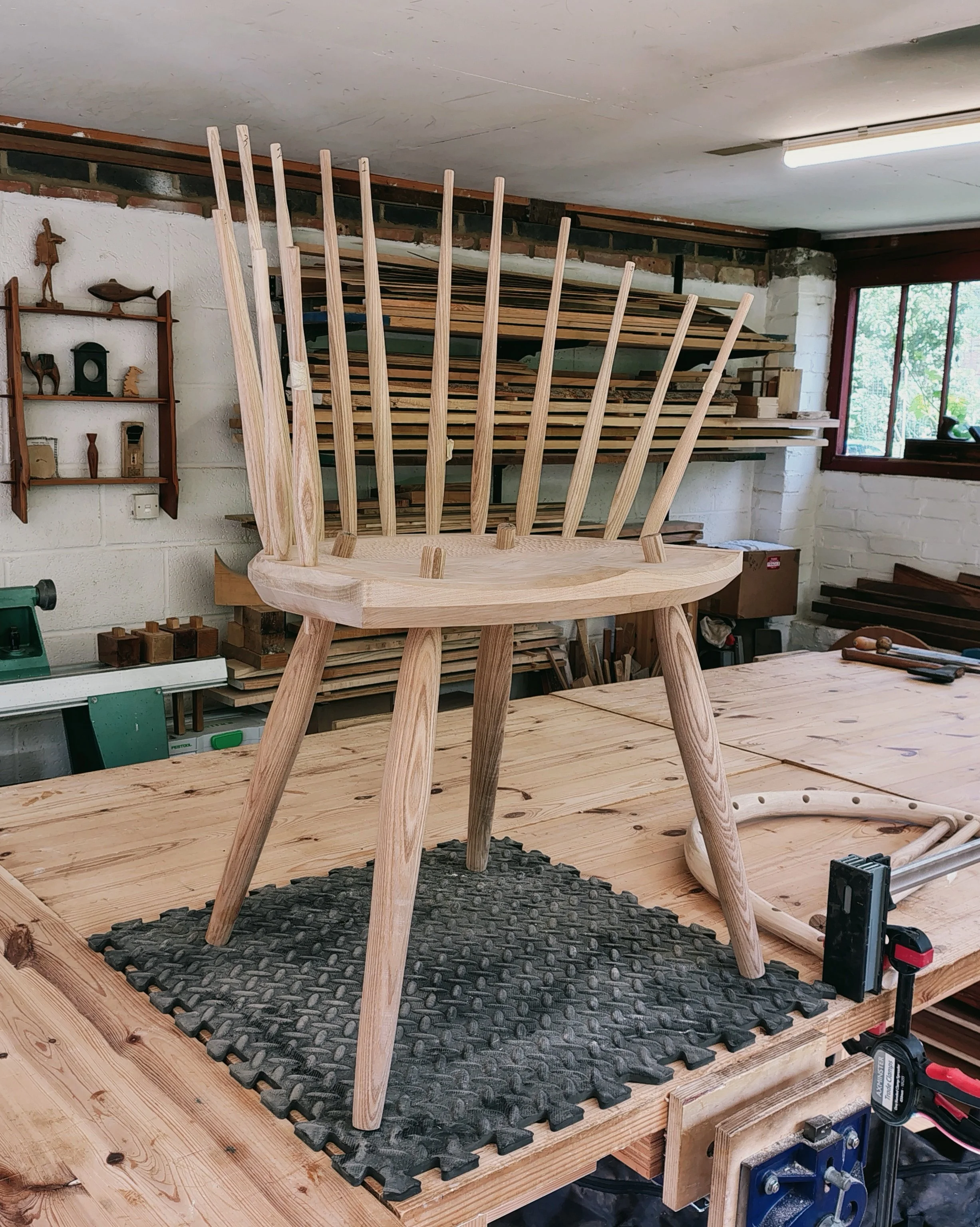 A wooden chair with natural wood finish sits on a black rubber mat on a woodworking workbench in a workshop. In the background, there are shelves with stacked wood planks, automotive tools, and a window letting in natural light.