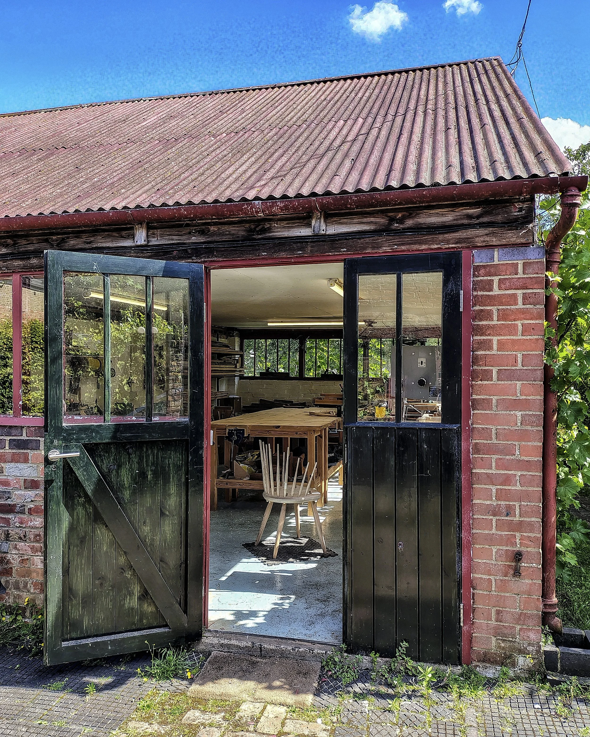 Open black door revealing a table and chair inside a brick-walled workshop or garage with a corrugated roof and window