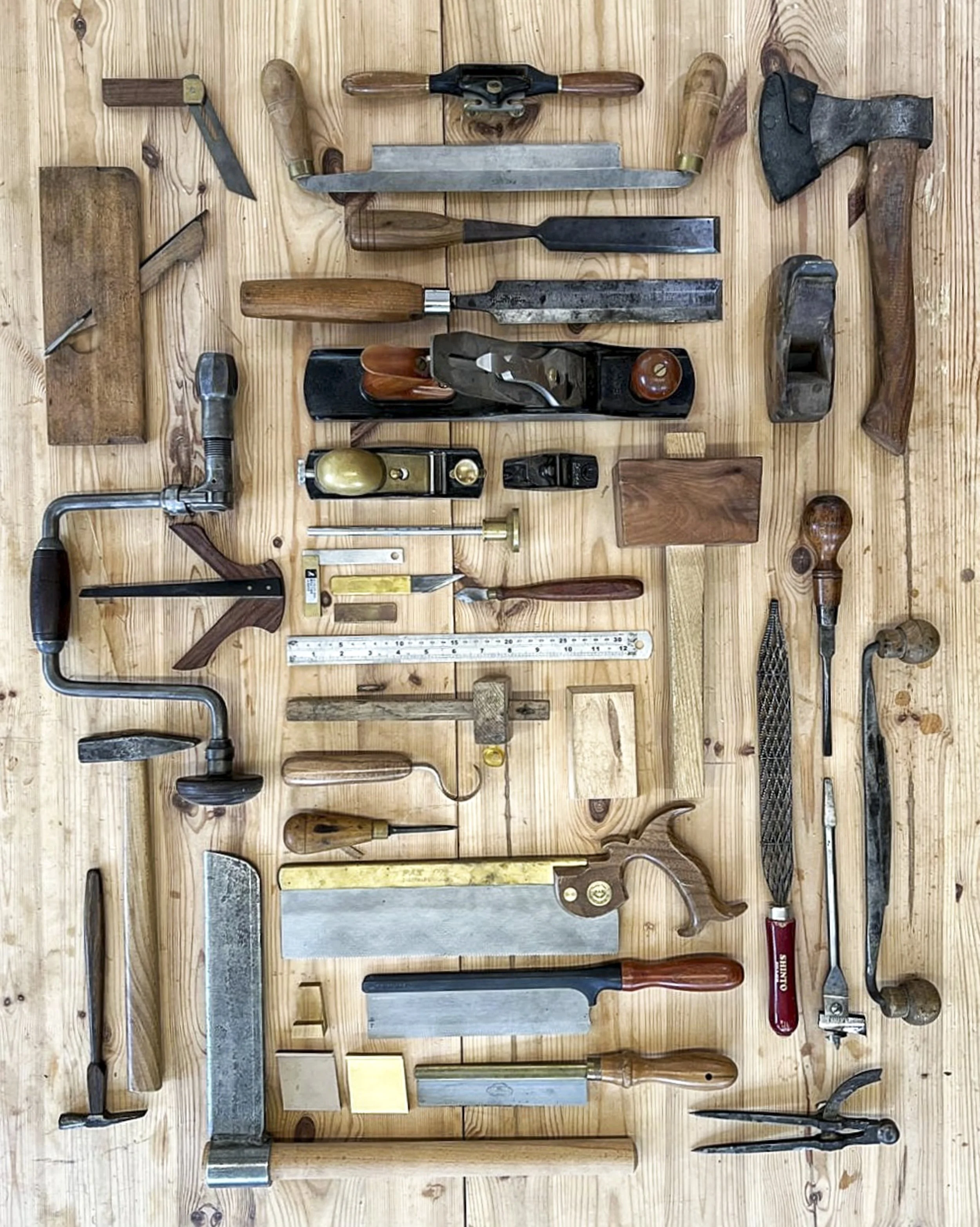 A collection of various hand tools and woodworking tools laid out on a wooden surface, including saws, hammers, chisels, a hand plane, a level, and a ruler.