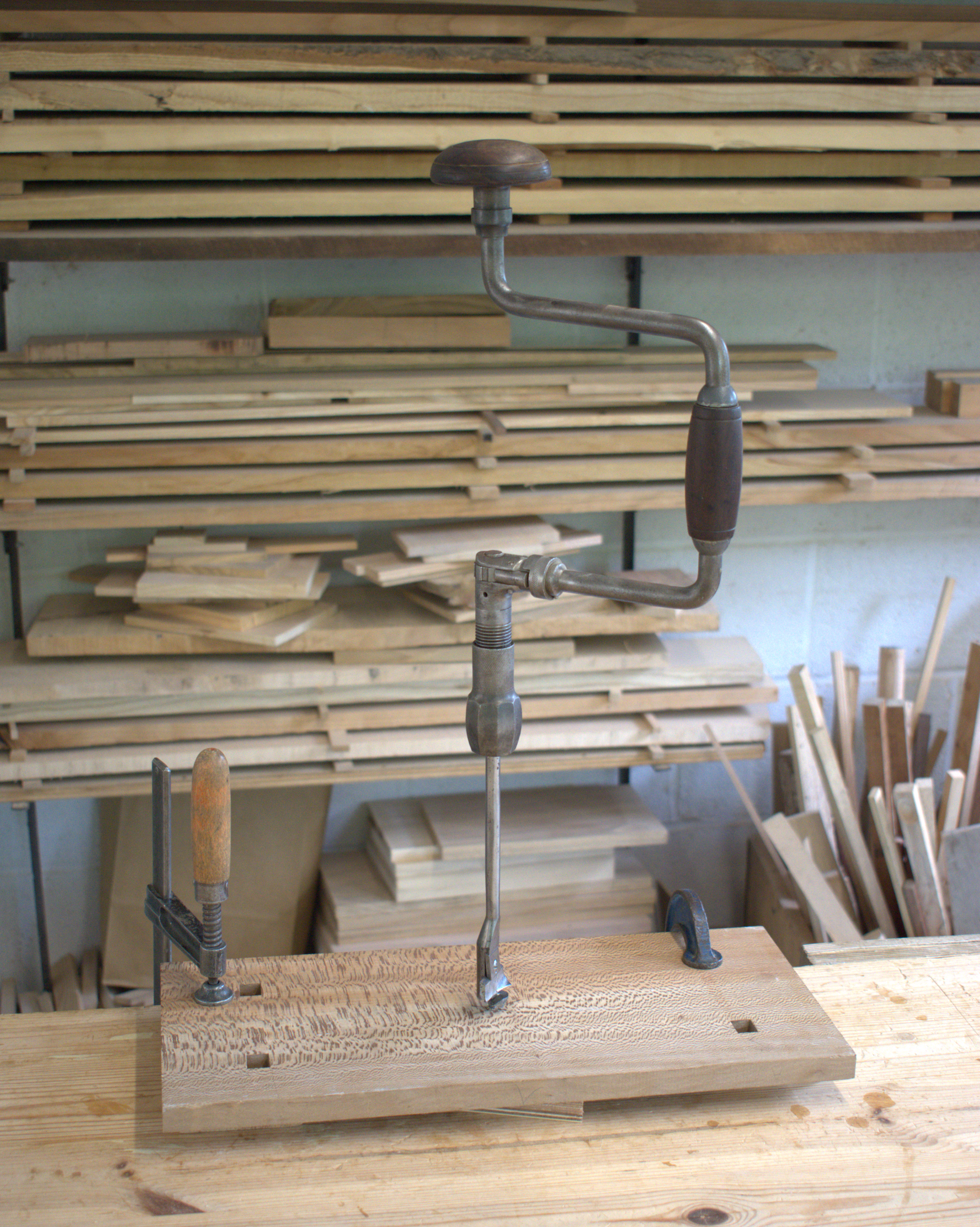 A woodworking clamp holding a piece of wood with a textured grain pattern on a workbench in a woodworking shop. In the background, wooden planks are stacked on shelves.
