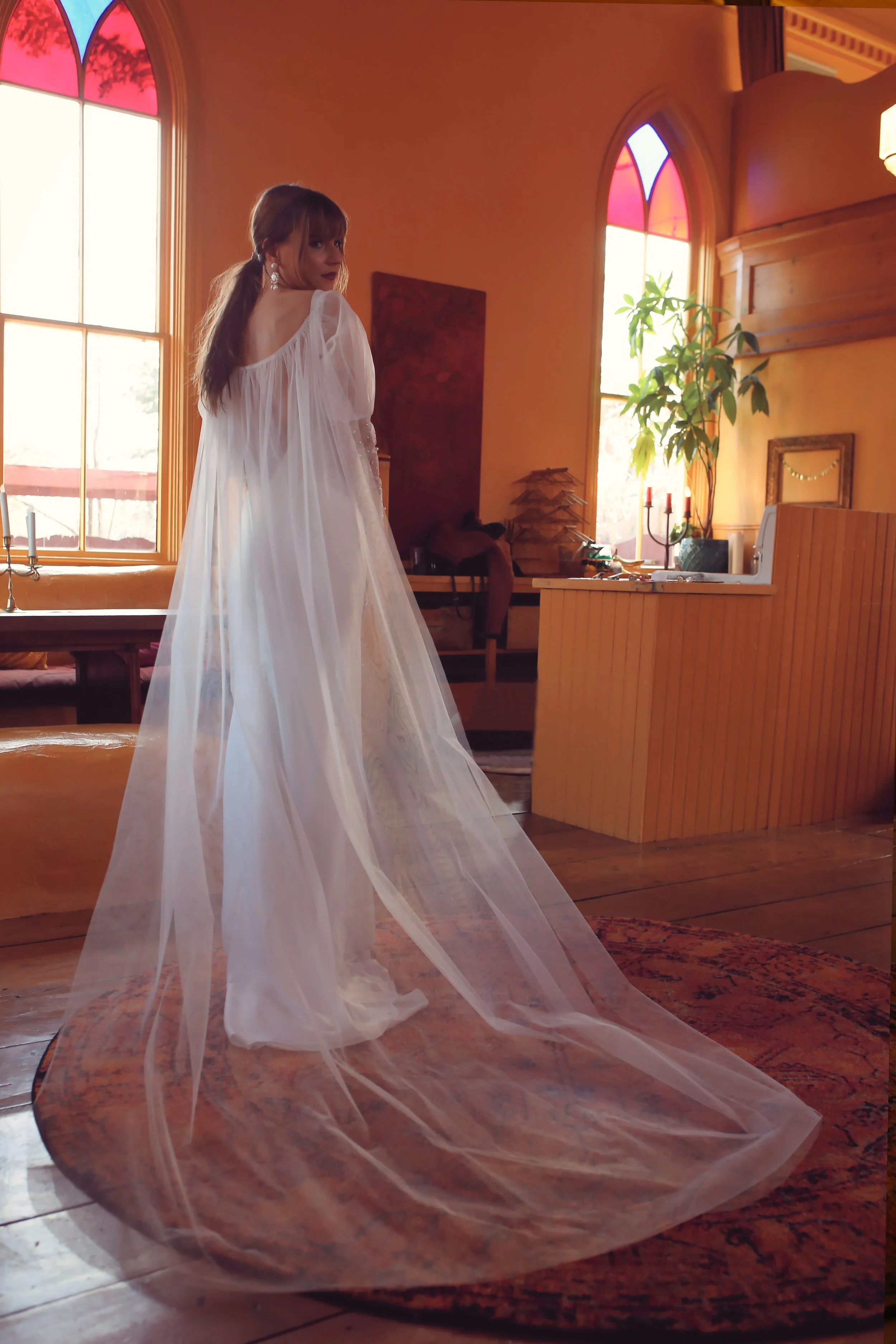 A woman in a flowing white dress with a long tulle train stands indoors near a window with stained glass. She has brown hair styled in loose waves and is wearing earrings, looking over her shoulder at the camera.