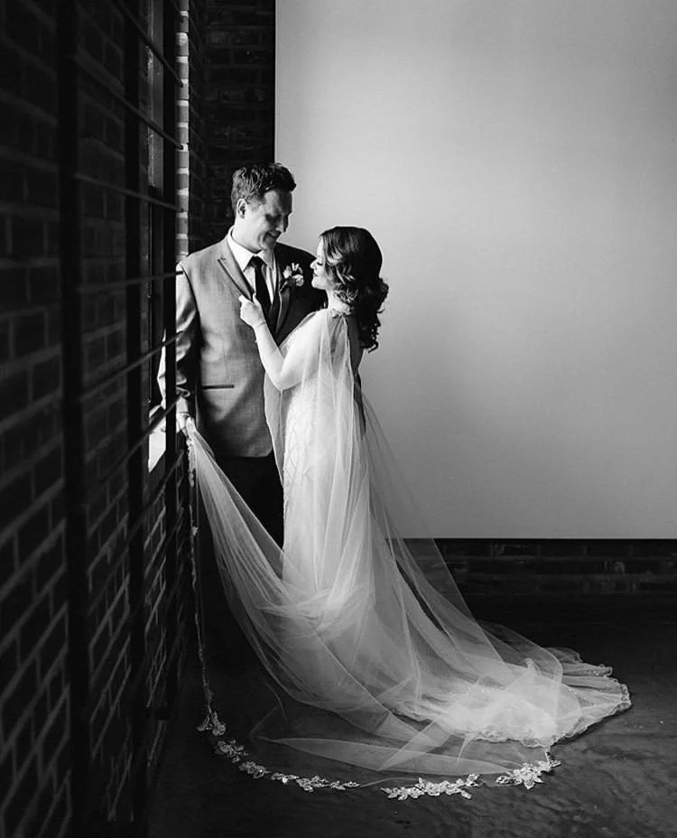 A bride and groom standing close together indoors, with the bride wearing a long veil and the groom in a suit. They are smiling at each other, standing next to a brick wall with a large window.