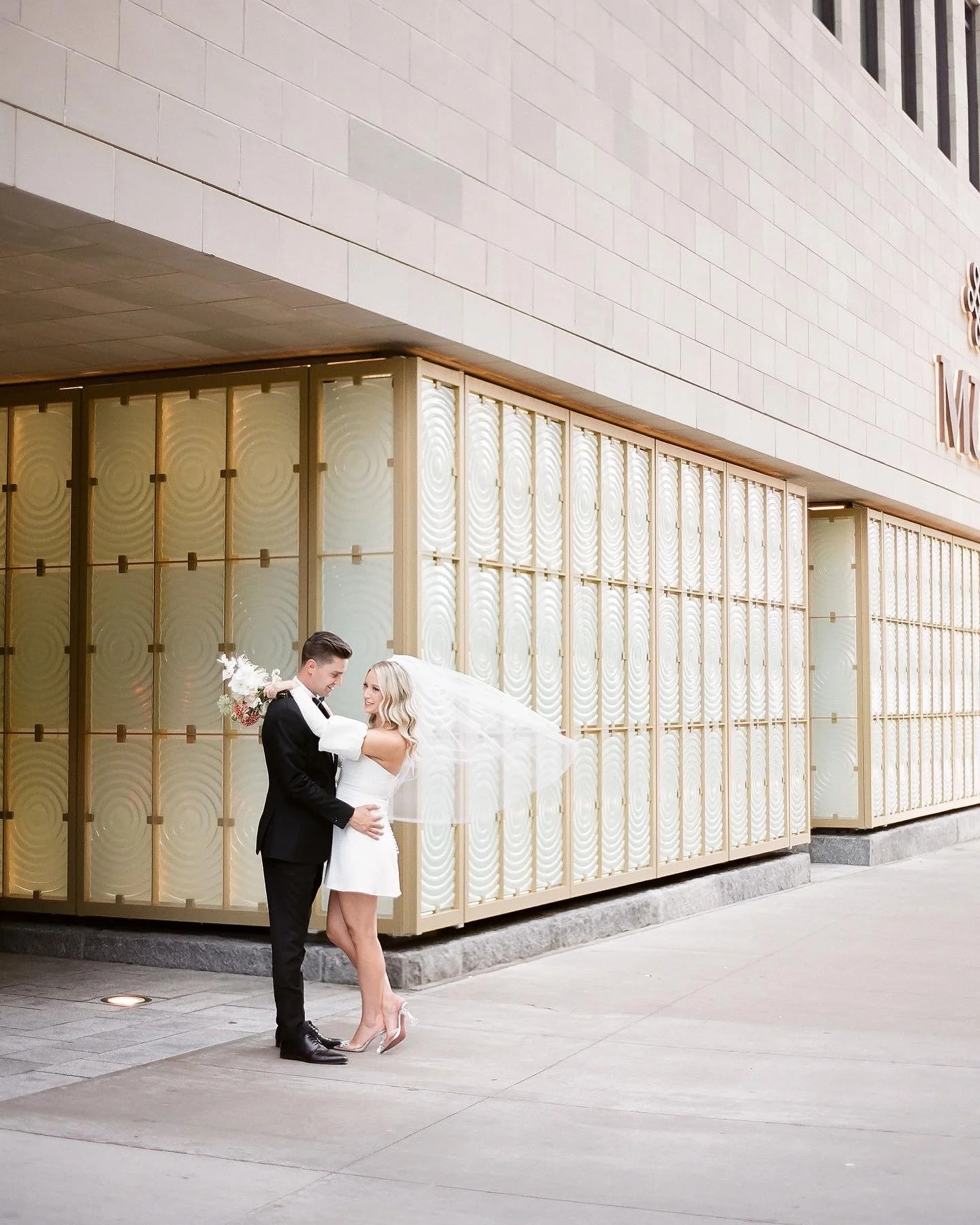 A newlywed couple standing outside a modern building with glass windows. The groom, dressed in a black tuxedo, is holding the bride, who is wearing a white dress and heels. The bride's veil is flowing in the wind, and they are smiling at each other.