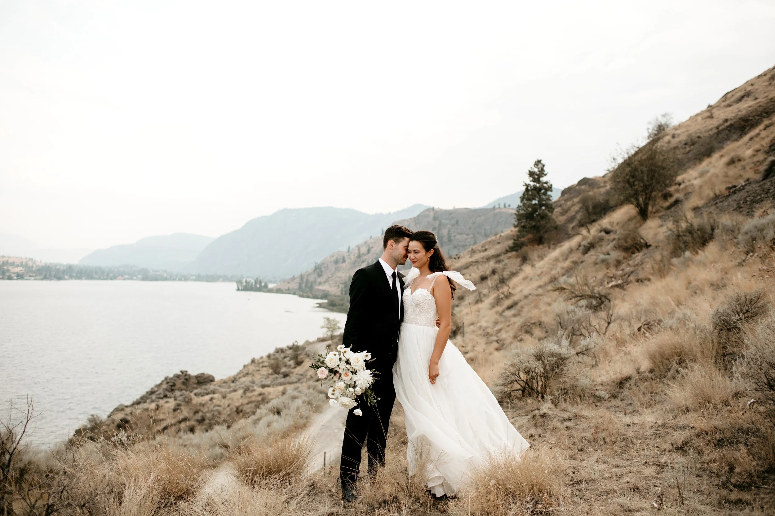 Wedding couple standing on a hillside by a lake with mountains in the background. The groom is wearing a black suit and the bride is in a white wedding dress holding a bouquet of white flowers.