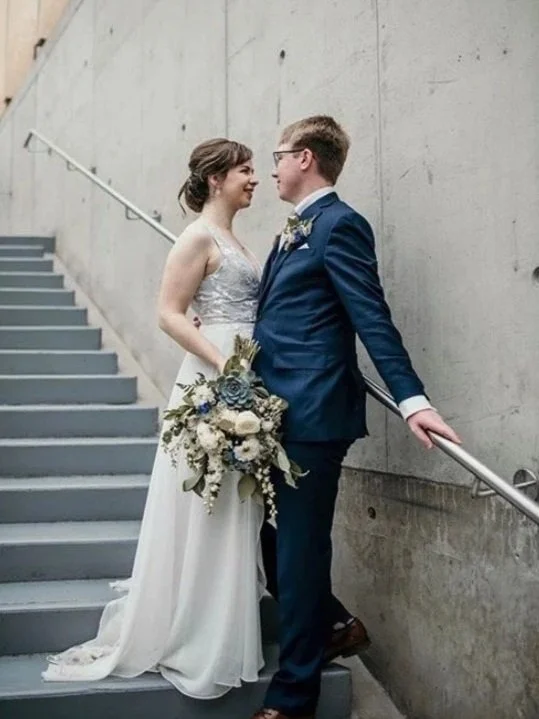 A bride and groom in wedding attire standing on a staircase, facing each other, with the bride holding a bouquet.