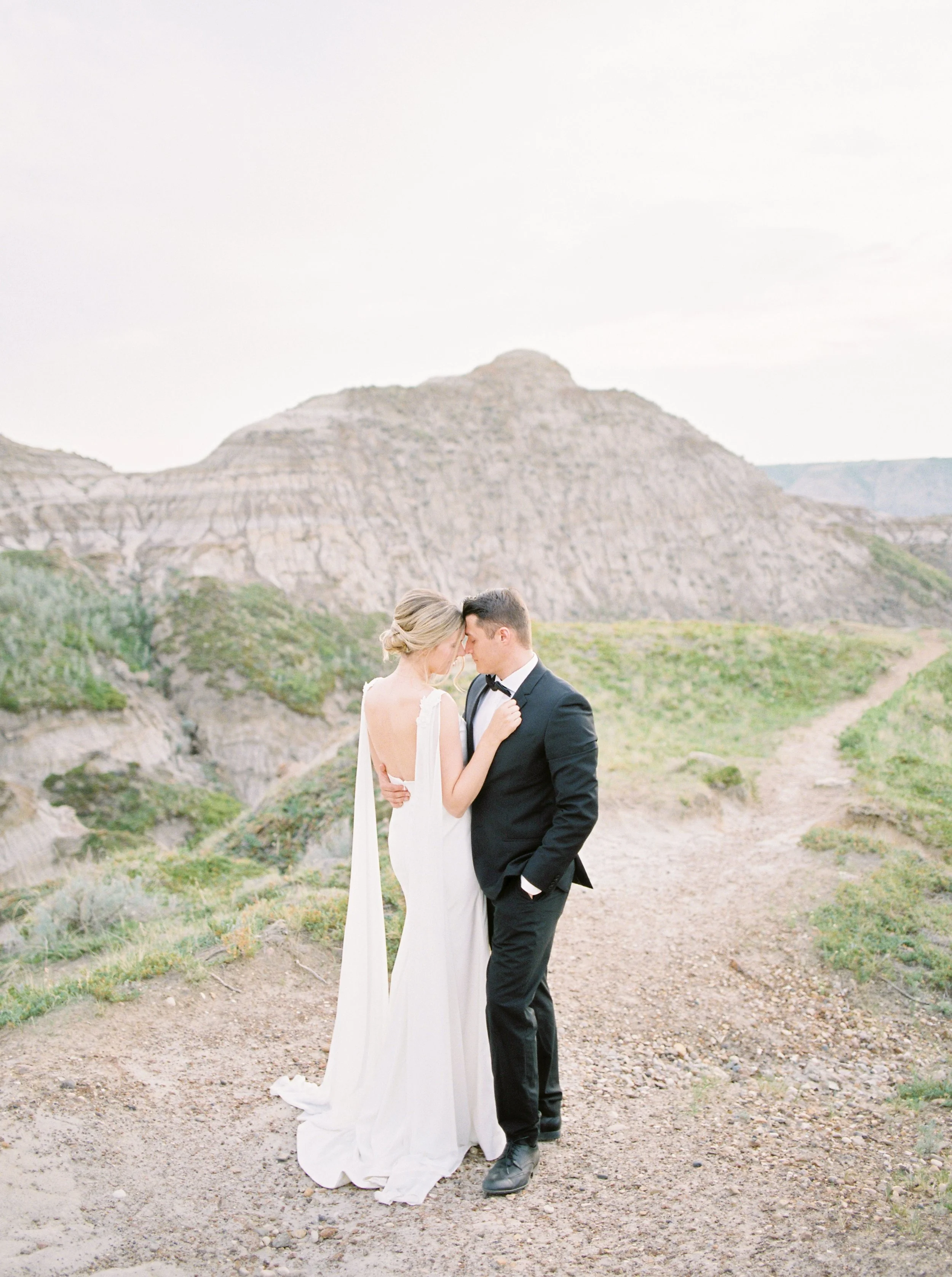 A bride and groom standing close together outdoors with mountains in the background. The bride is wearing a white wedding dress and the groom is wearing a tuxedo. They are touching foreheads with eyes closed, sharing an intimate moment.