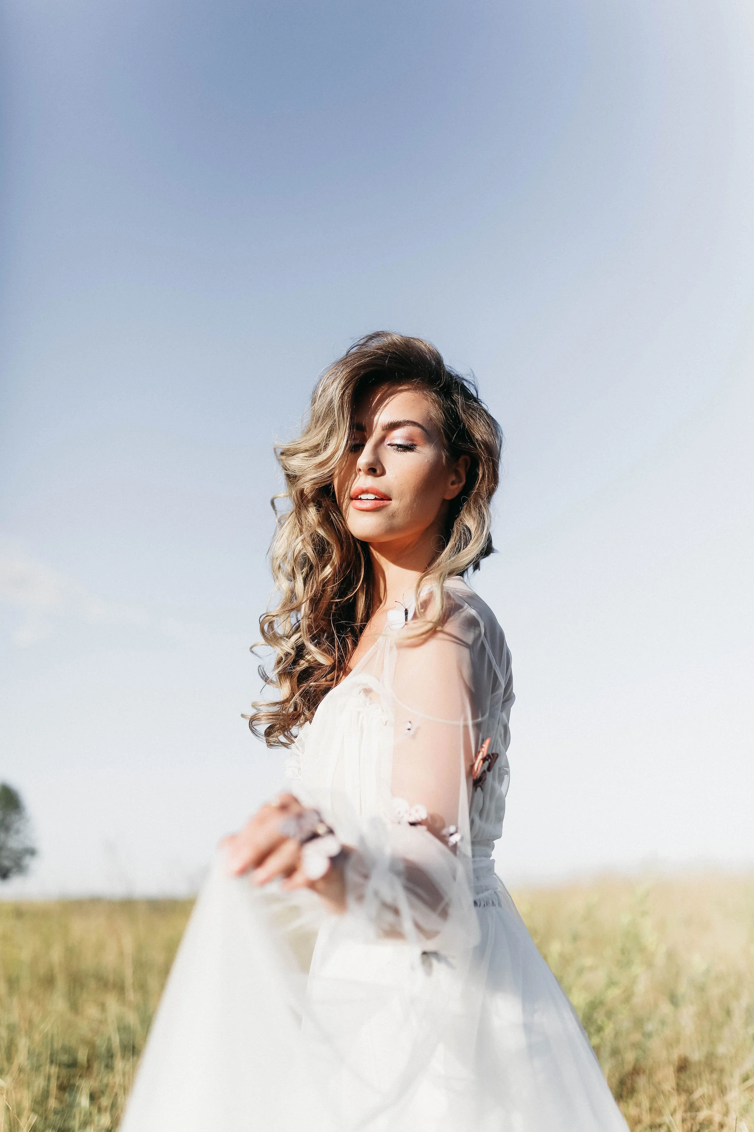 A woman with long wavy hair wearing a white dress standing in a field with her eyes closed and a slight smile, with a clear blue sky in the background.
