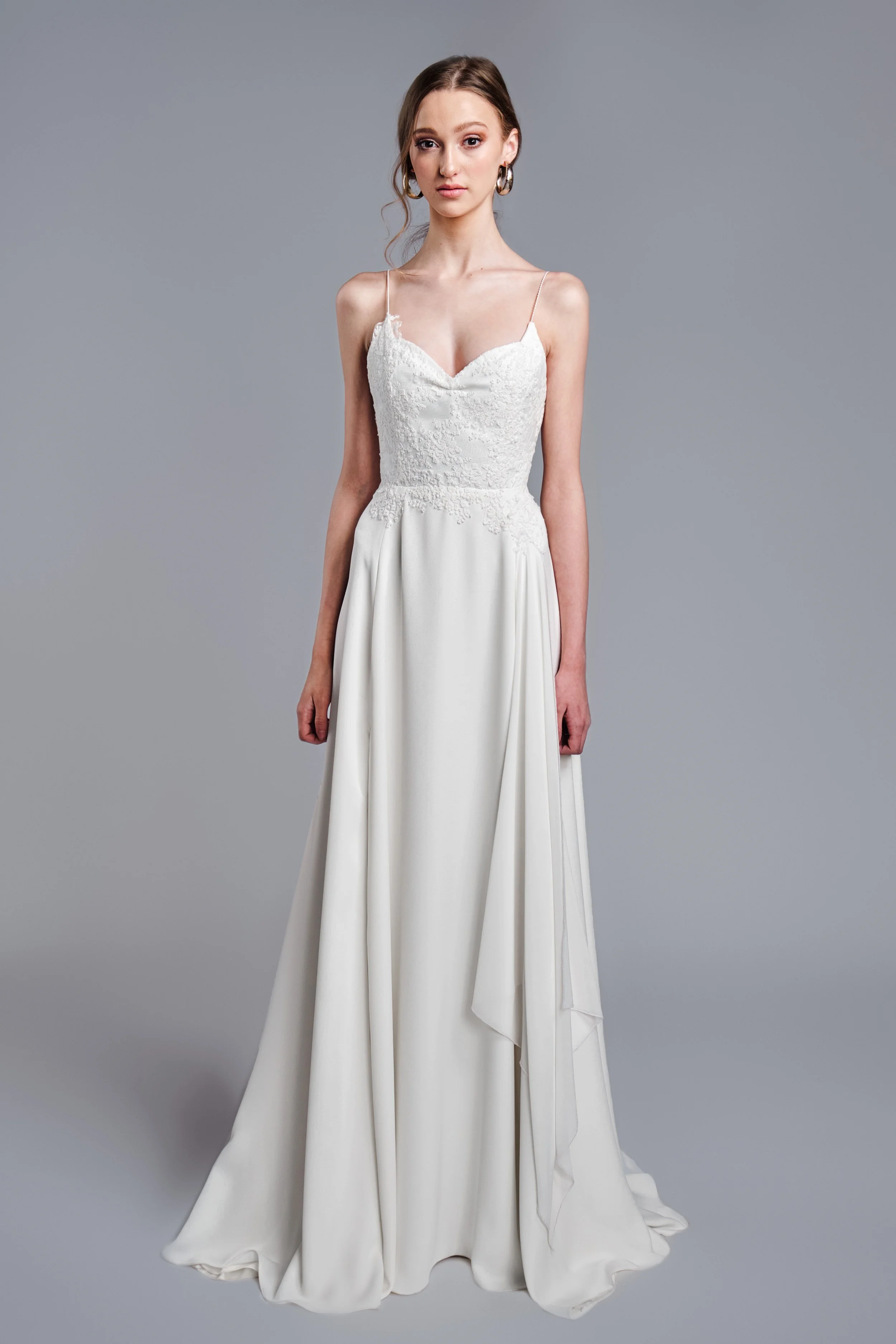 A woman in a white wedding dress with lace details on the bodice, thin spaghetti straps, and a flowing skirt stands against a plain gray background, looking directly at the camera.