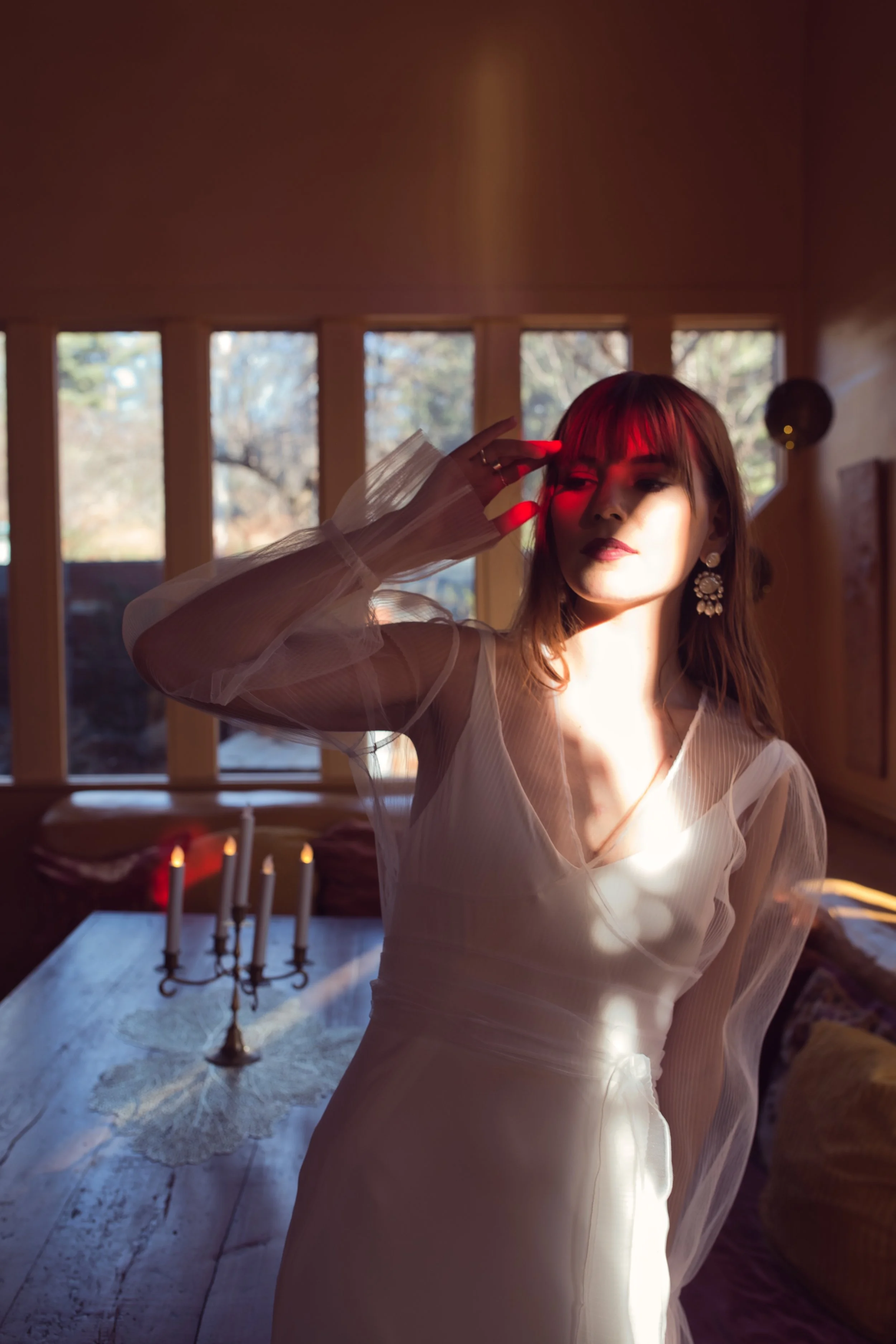 A woman with shoulder-length brown hair and large floral earrings wearing a sheer white dress with puffed sleeves, standing in a wooden room with large windows and natural light. There is a candelabra with five lit candles on a wooden table and a pur