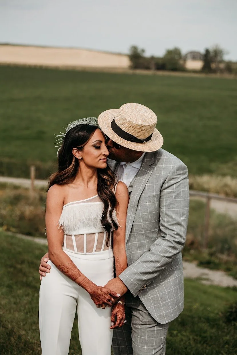 A couple in wedding attire stands in a green outdoor setting, with the man wearing a gray checkered suit and straw hat, and the woman wearing a white dress with fringe and intricate henna designs on her arms, holding hands closely.