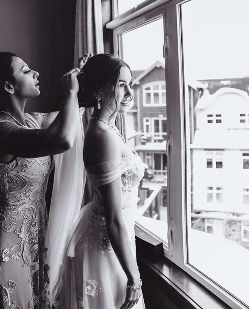A bride standing by a window with her hair being styled by a woman. The bride is wearing a wedding dress and jewelry, and looks out the window with a smile. The scene is in black and white.