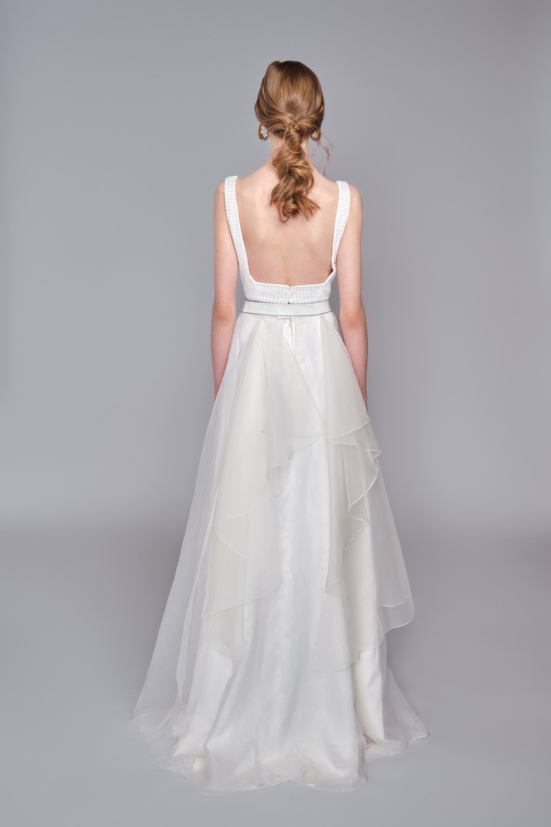 Back view of a woman wearing a white sleeveless dress with tiered, flowing skirt and a low back, standing against a plain gray background.