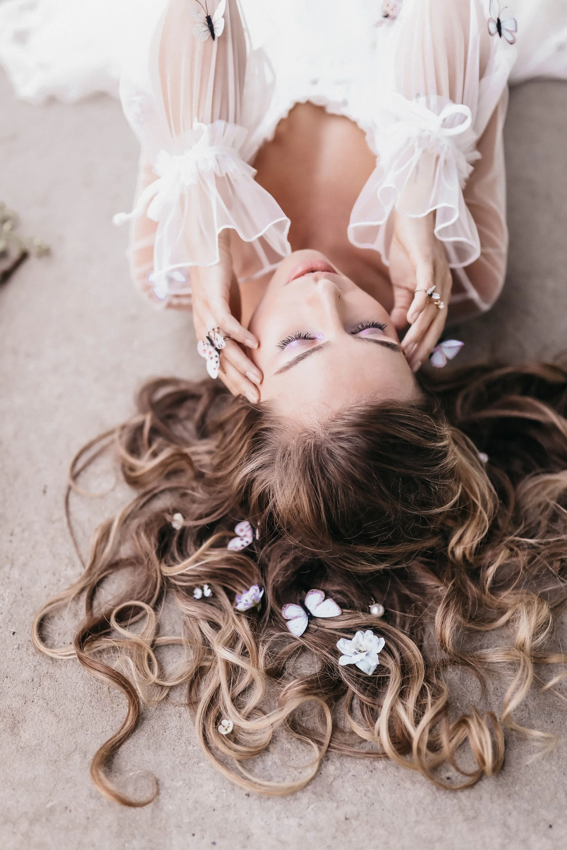 A woman with long, curly blonde hair lying on a sandy surface. She has her eyes closed, wearing a light, ruffled blouse with butterflies on the sleeves, and has flowers and butterflies in her hair.