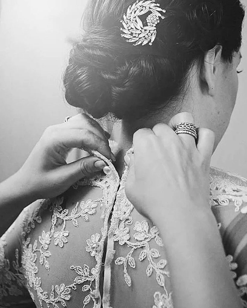 A woman is adjusting her lace wedding dress, with her hair styled in an elegant updo adorned with a decorative hairpiece, and wearing rings on her fingers.