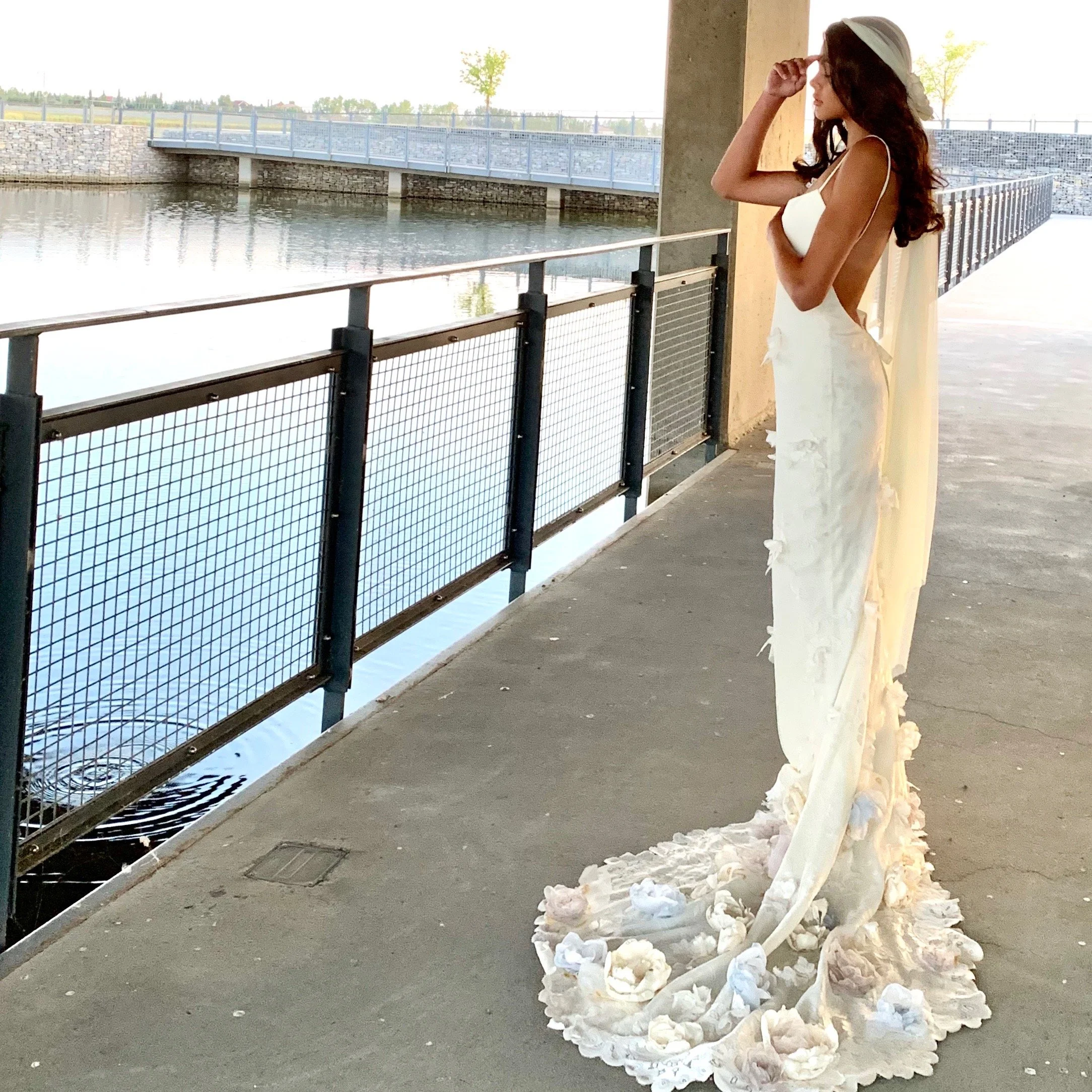 A woman in a white dress and hat standing on a riverside promenade, looking out over the water with her hand adjusting her hat.