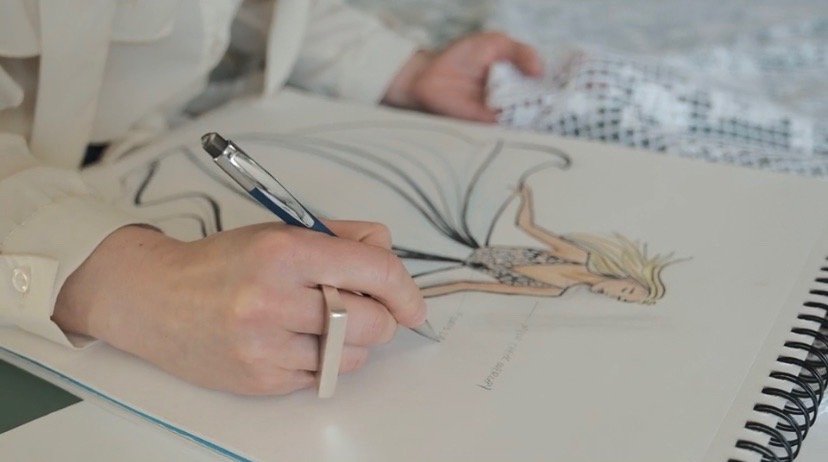A person drawing a fashion illustration of a woman in a dress with a pen on an art sketchpad, with a hospital bracelet visible on their wrist.