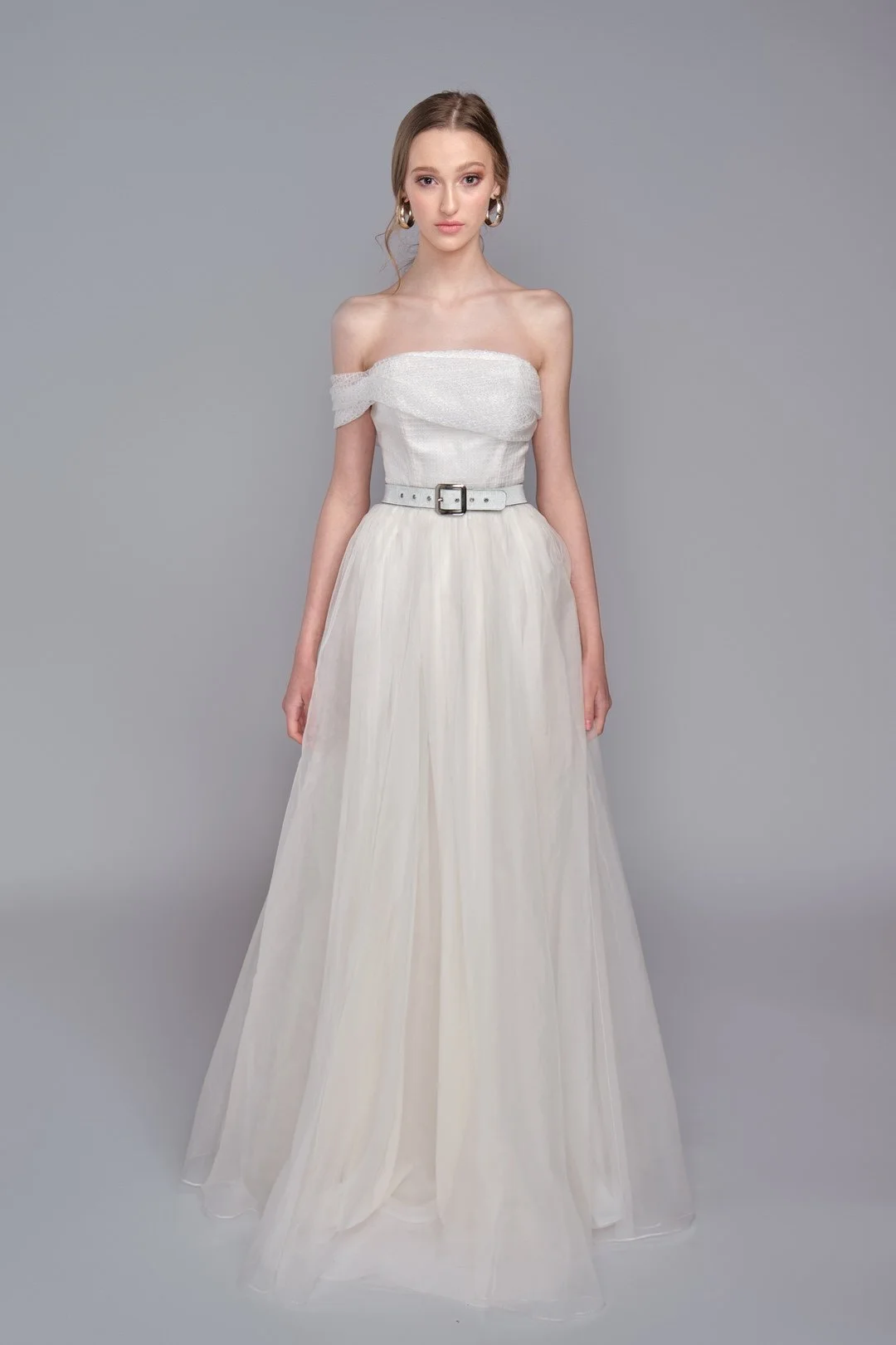 A young woman in an off-shoulder white gown with a belt, standing against a plain gray background.