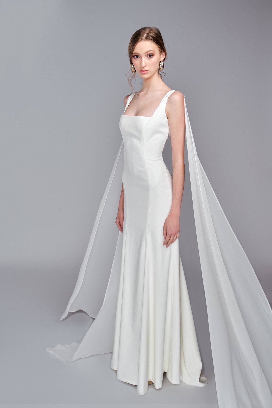 A young woman with brown hair in a loose bun, wearing a white satin wedding gown with a square neckline, fitted bodice, and a long train with a cape-like extension over her shoulders, standing against a plain gray background.