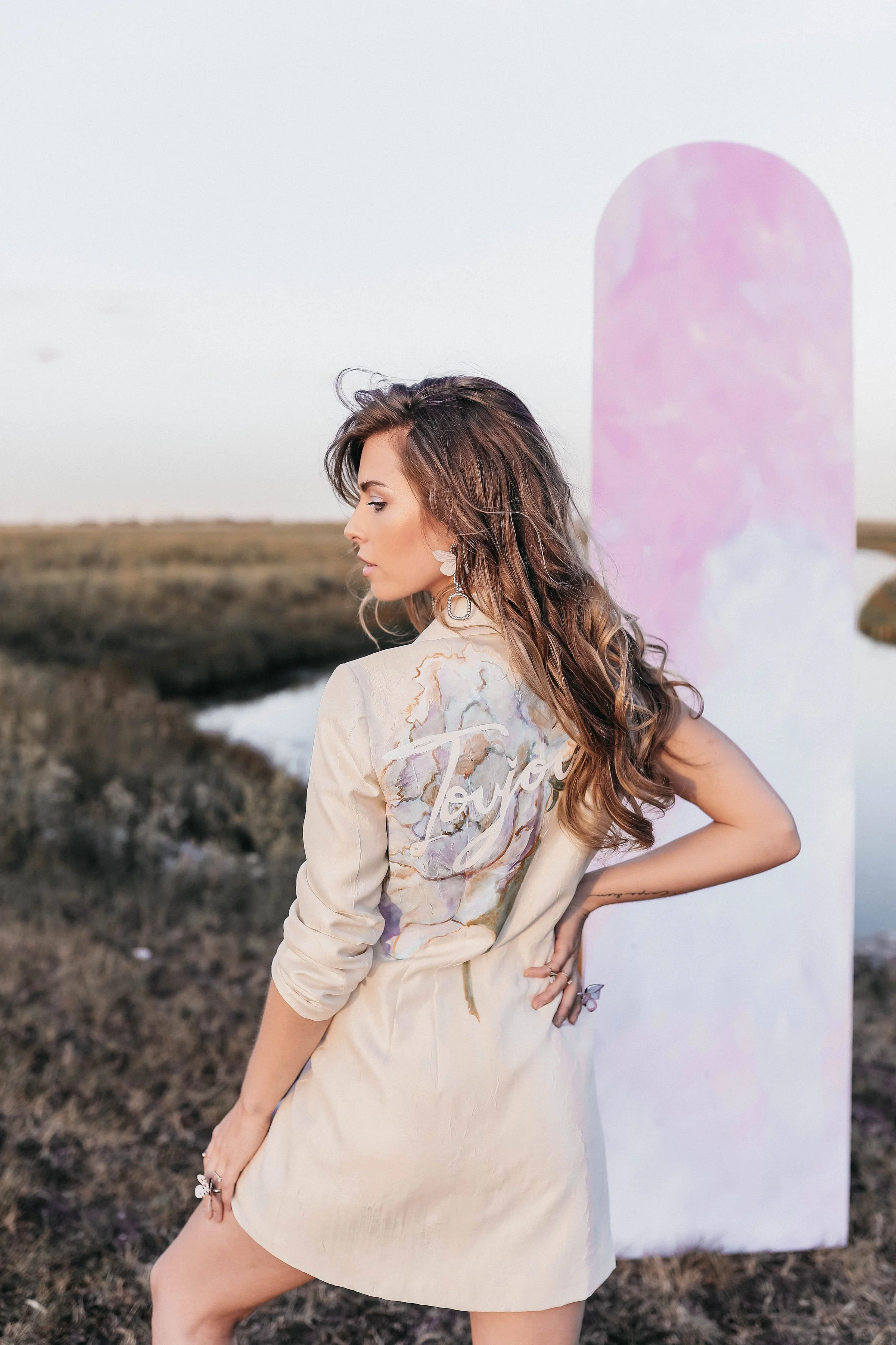 A woman with wavy brown hair wearing a beige dress with floral patterns and the word 'Taylor' written on it, standing outdoors near a water body with marshy land, her right hand on her hip, and large earrings, with a pastel pink and purple abstract b