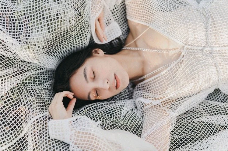 A woman with dark hair sleeping peacefully on a white, mesh-covered surface, wearing a light-colored, see-through garment.