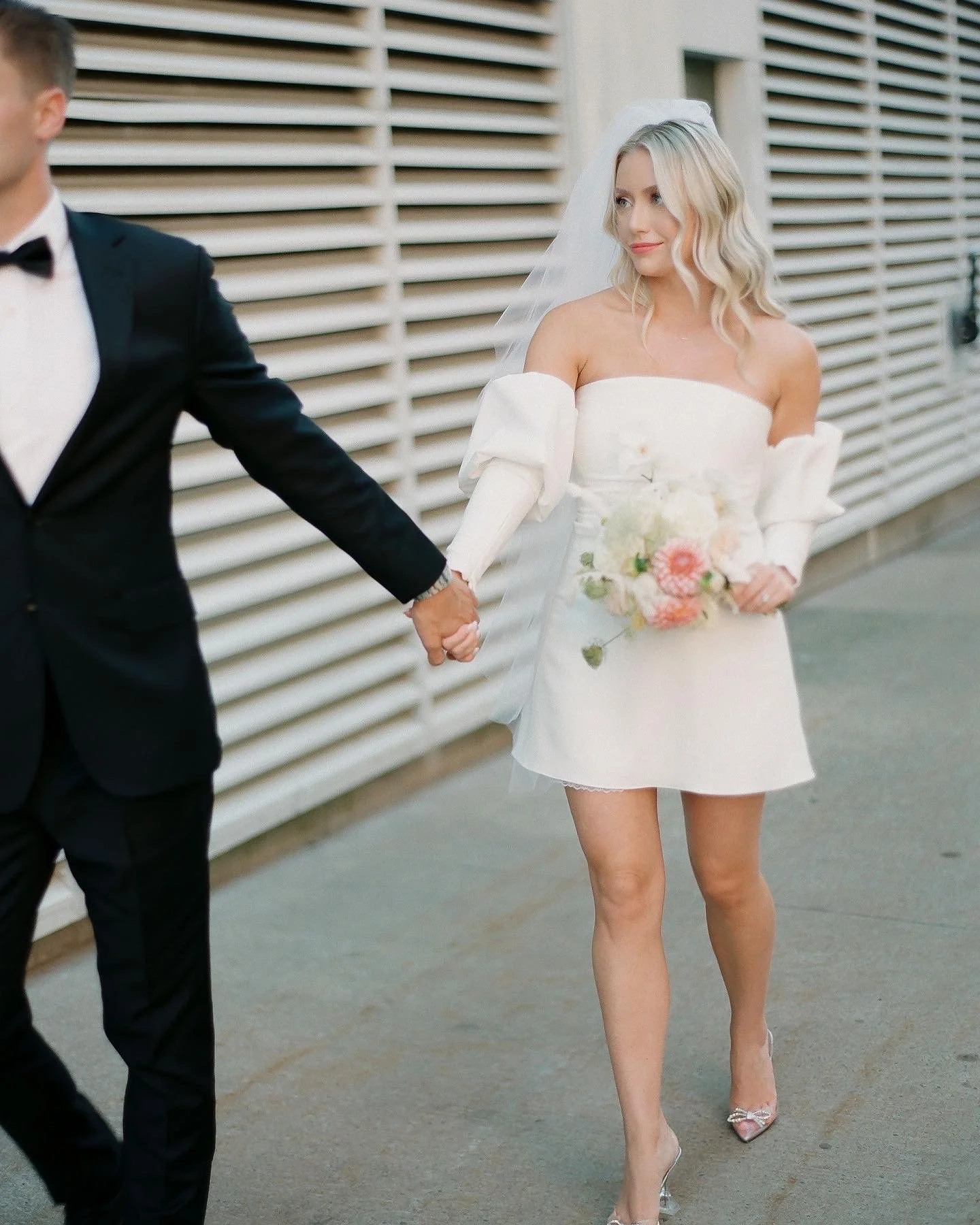 A woman in a white off-the-shoulder dress holding a bouquet of flowers, walking hand-in-hand with a man in a black tuxedo, against a background of horizontal white slats.