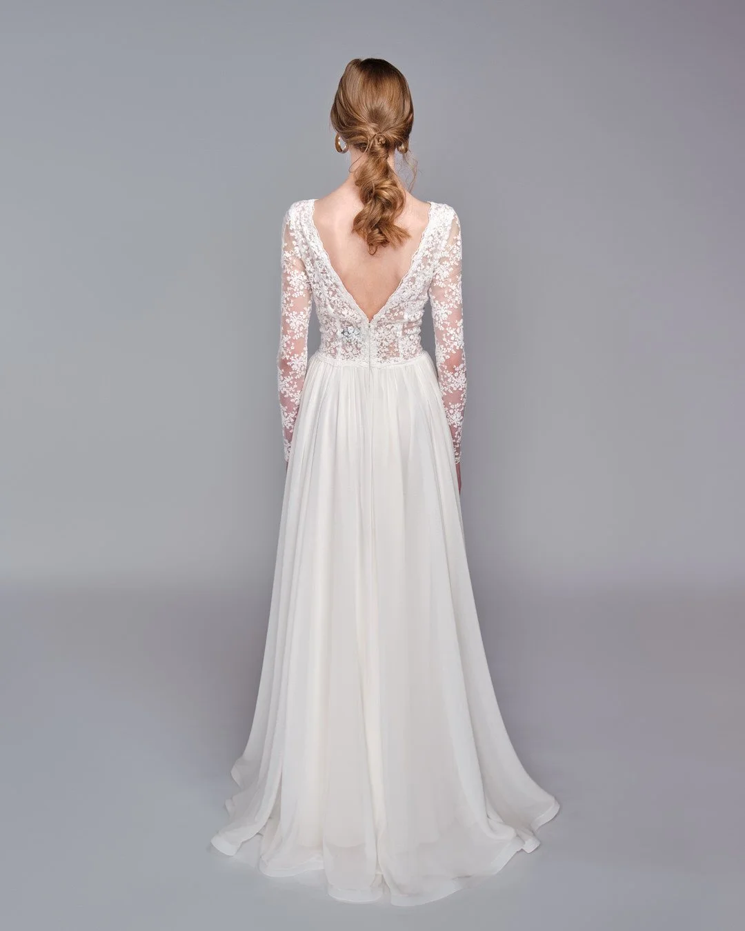 Back view of a woman in a white lace long-sleeve wedding dress with a deep V back, standing against a plain gray background.