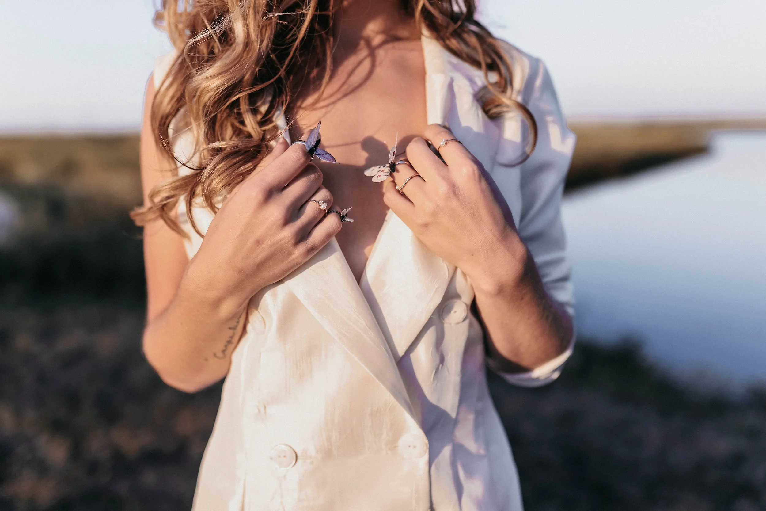 Close-up of a woman with curly hair wearing a cream-colored blazer, holding butterfly-shaped rings on her fingers, outdoors with a blurred natural background.