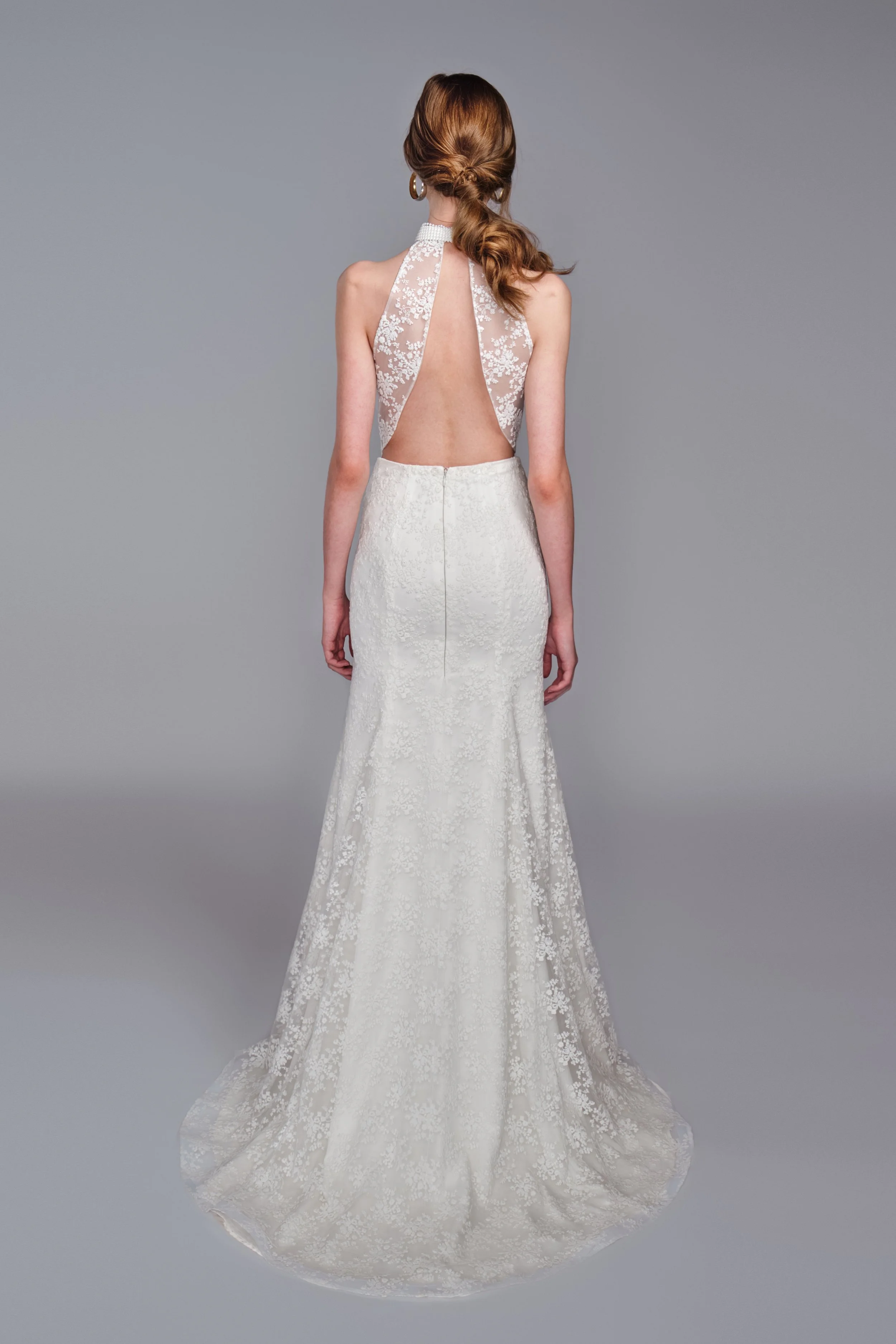 Back view of a woman in a white wedding gown with lace details, featuring a low-cut open back, standing against a plain gray background.