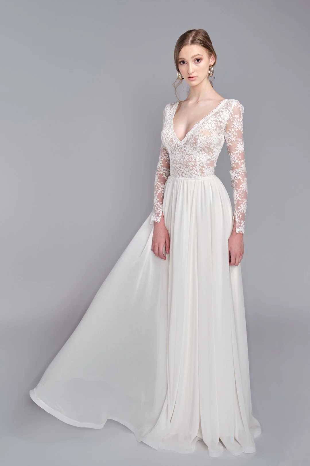 A woman in a white wedding dress with a lace bodice and long sleeves, standing against a plain gray background.