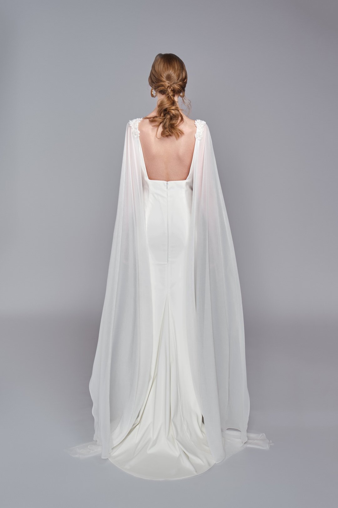 Back view of a woman wearing a long white wedding dress with cape-style sleeves and floral embroidery on the shoulders, standing against a plain gray background.