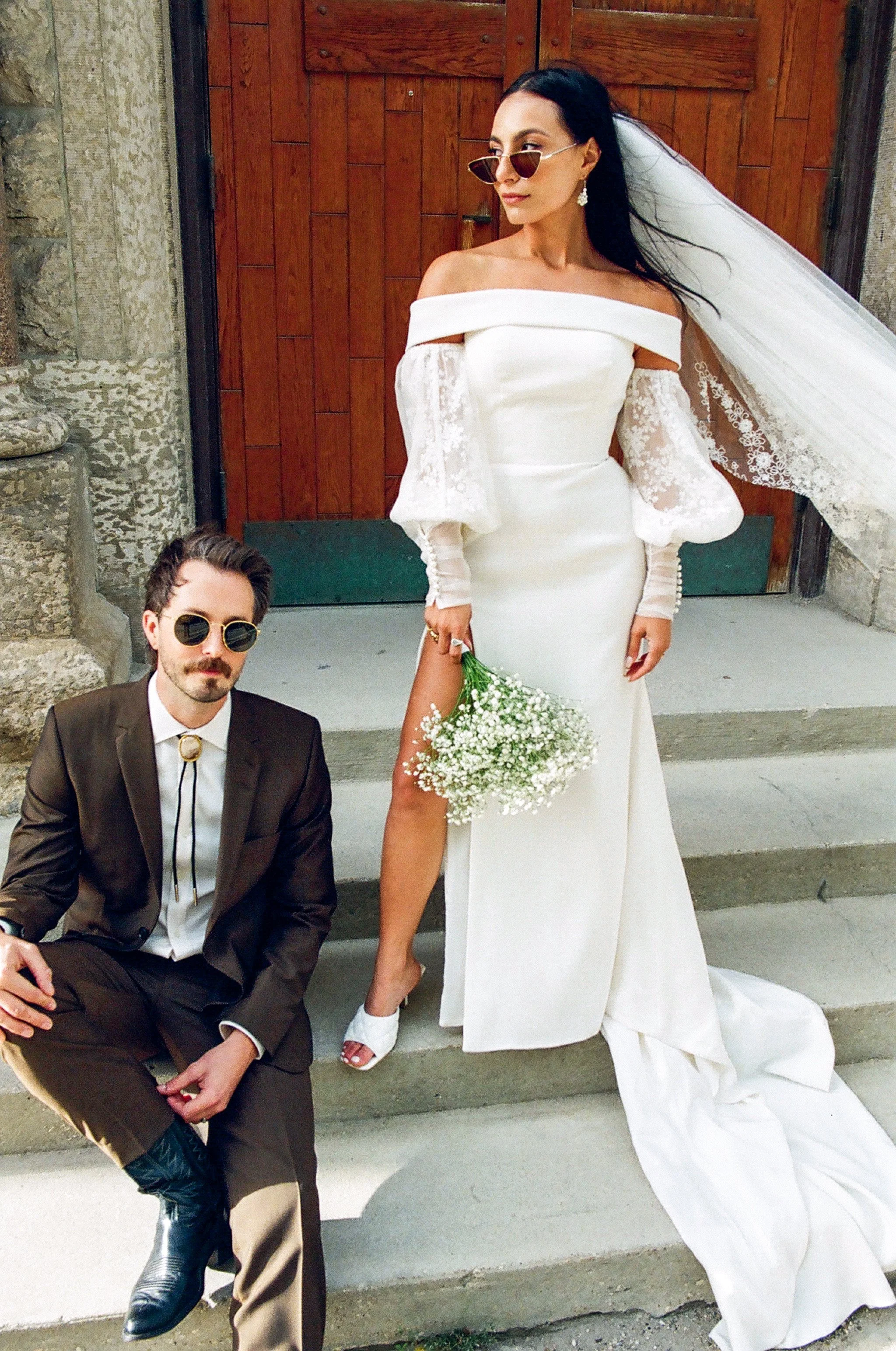 A woman in a white wedding dress with off-the-shoulder sleeves and lace detail, holding a bouquet of white flowers, standing next to a man in a dark suit with a bolo tie and sunglasses, sitting on stairs outside a stone building with wooden doors.