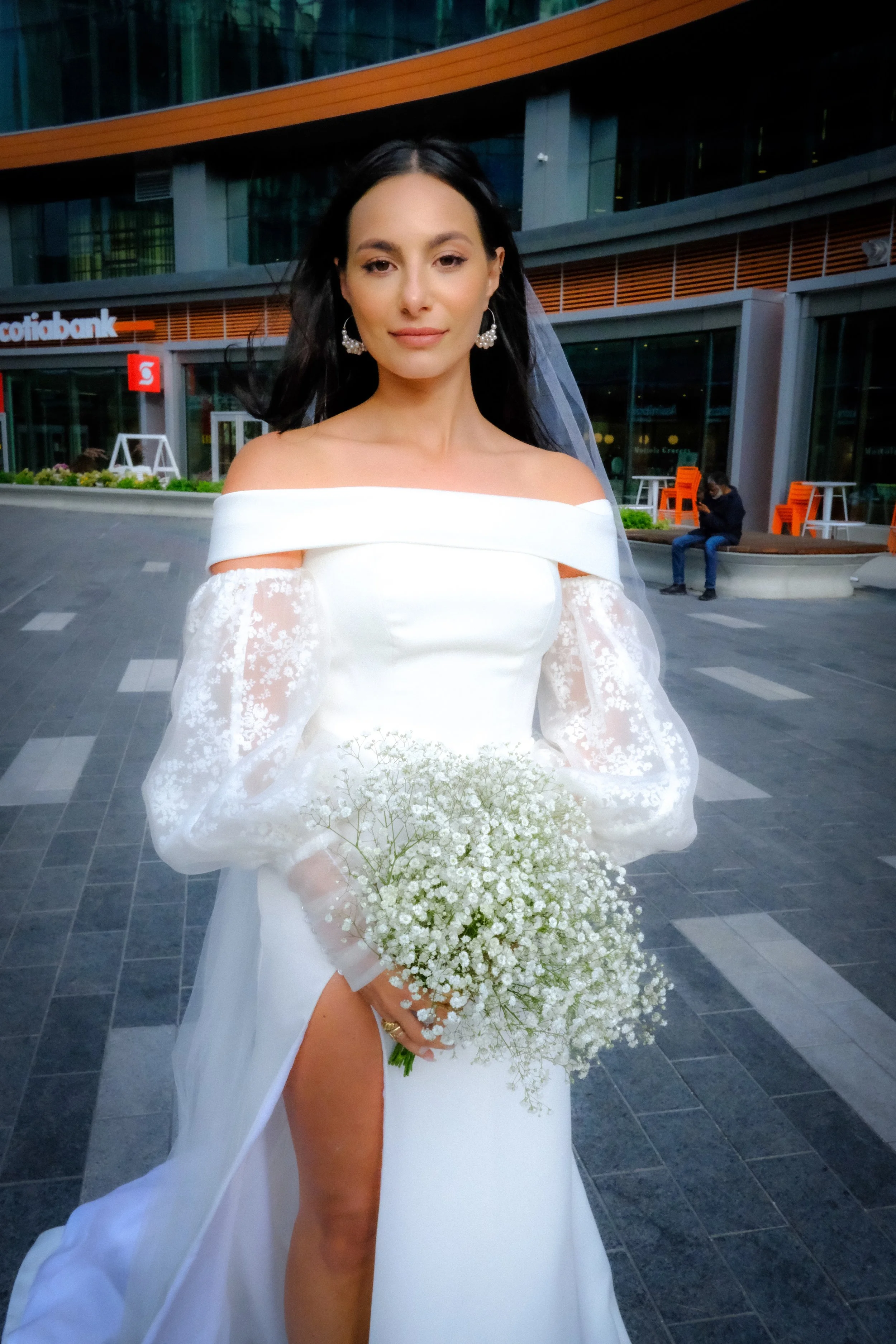 A woman wearing a white off-shoulder wedding dress with lace sleeves, holding a bouquet of small white flowers, standing outside a modern building with orange and green accents.