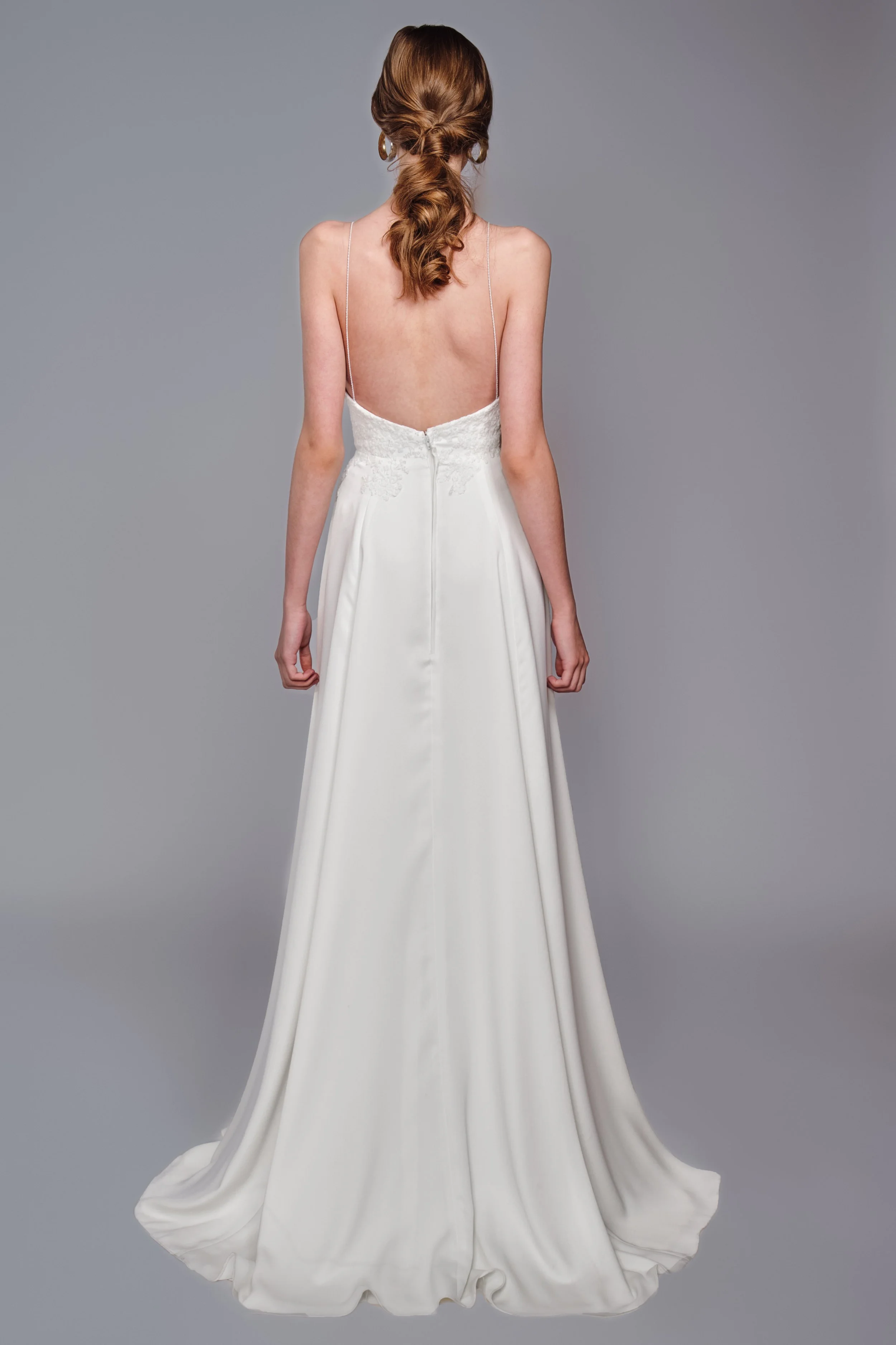 Back view of a woman with long, wavy, red hair in a loose ponytail, wearing a white, strapless gown with thin spaghetti straps and a zipper closure at the back, standing against a plain gray background.