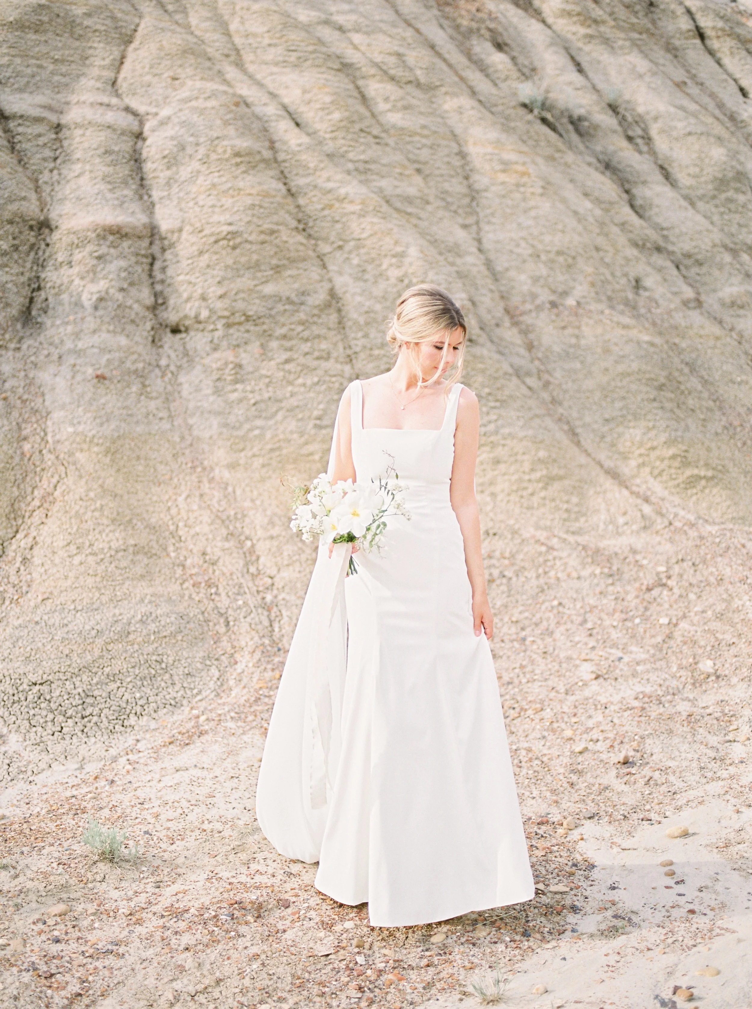 A woman in a white wedding dress holding a bouquet of white flowers standing on a rocky, desert-like terrain with steep, sandy cliffs in the background.