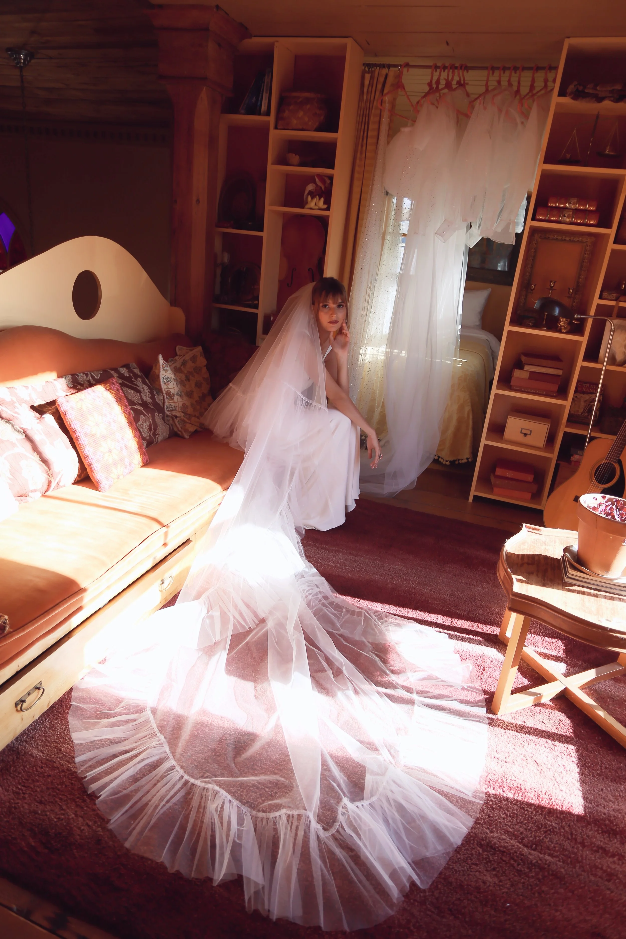 A woman dressed as a bride sitting on a sofa in a room with wooden walls. She is wearing a white wedding dress with a long, flowing veil. The room has a bookshelf, a bed, and a window with sheer curtains, with dresses hanging on a rod.